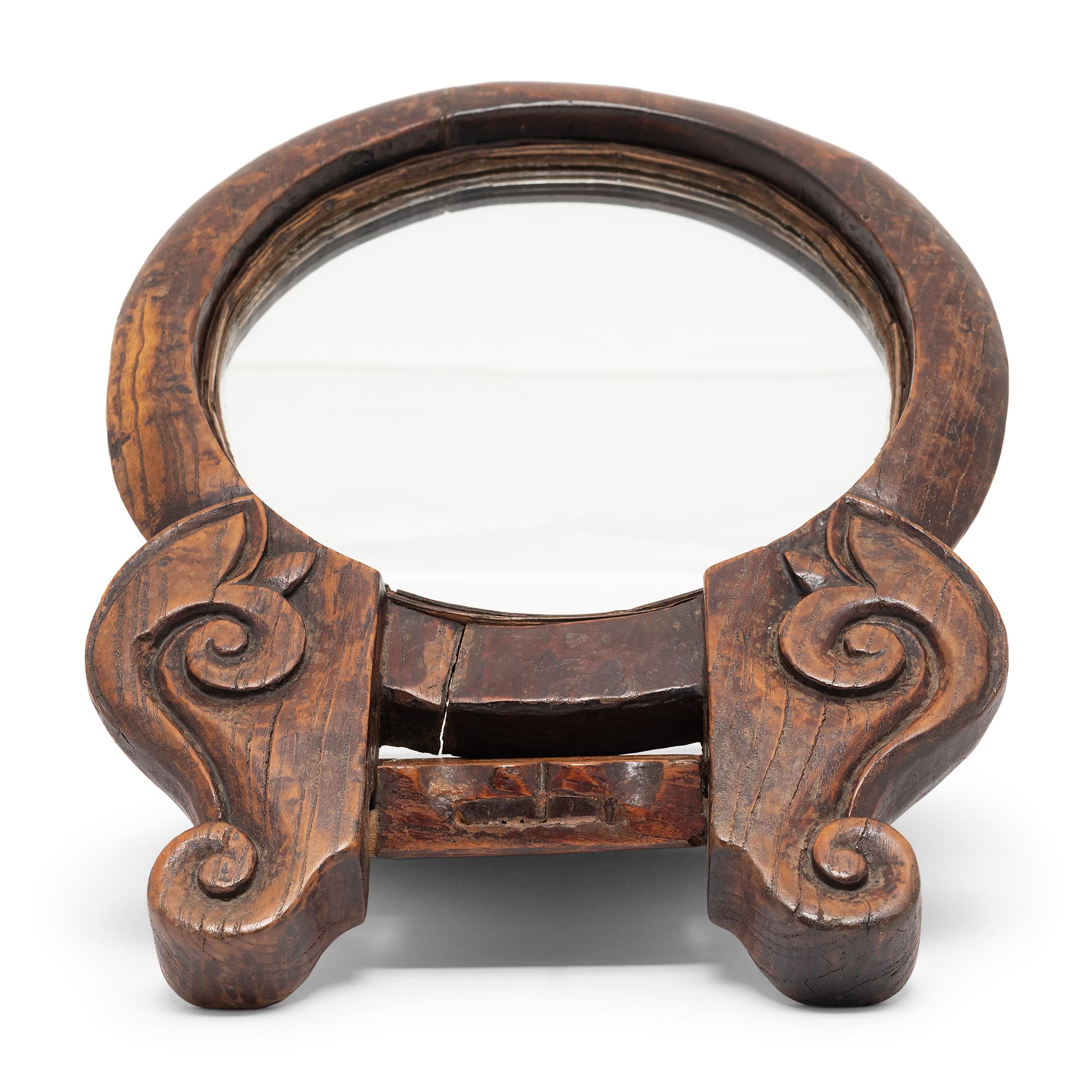 This round folding mirror dates to the mid-19th century and would have accompanied a well-to-do woman on her travels. The wooden frame is carved with simple scrollwork at the hinge and includes a flat cover that props the mirror upright when open.