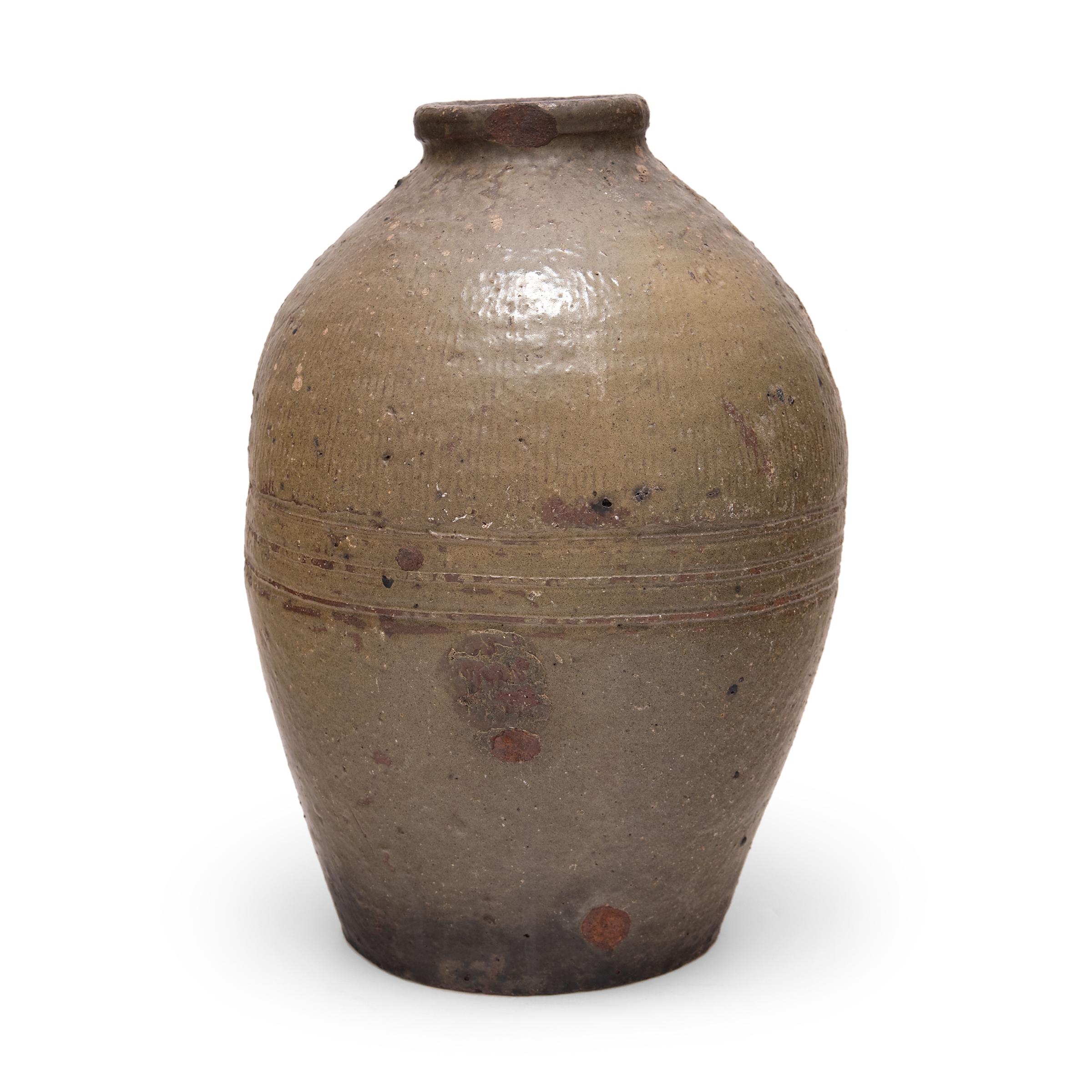 Originally used as a fermenting jar, this early 20th century ceramic pot is coated inside and out with a subdued green-brown glaze. The jar has a narrow mouth and a rounded, oblong form that tapers to the base. Shaped by paddling, the jar has a