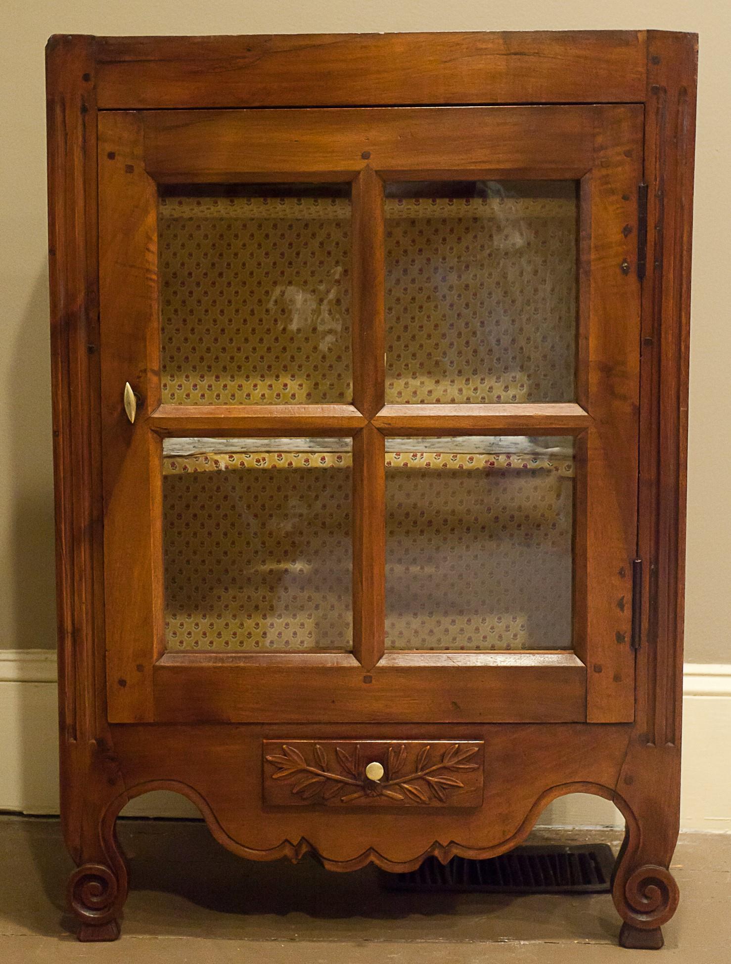 This small glass-door cabinet has short cabriole legs so it can be used as a standing piece, but it also has original cast iron hooks on its sides for use as a hanging cabinet possibly on shipboard. Made of fruitwood. Door has 4 old glass panes. One