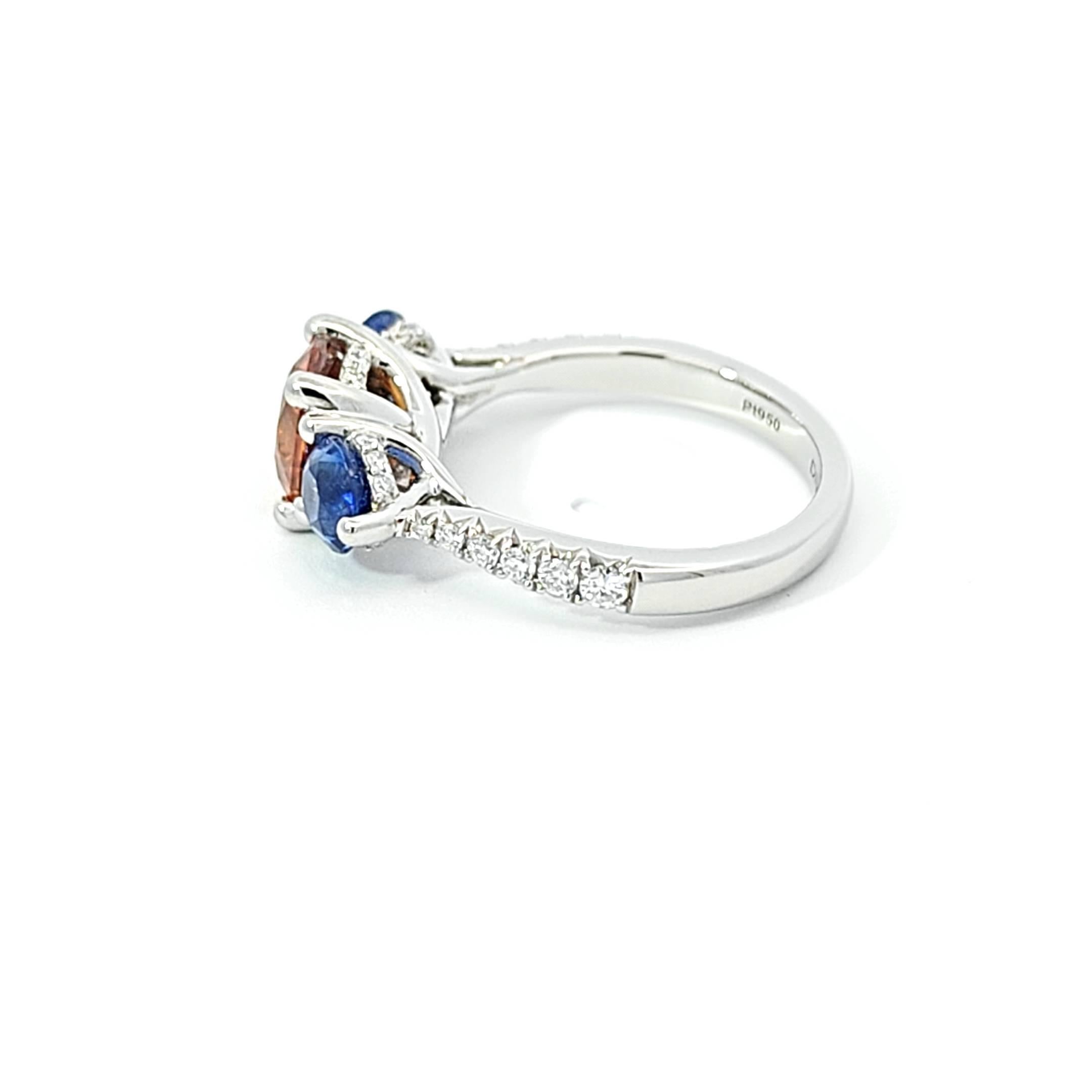 This exquisite platinum ring celebrates a kaleidoscope of colors with its vibrant gemstone centerpiece, featuring a fiery orange zircon nestled between two rich blue sapphires. Crafted to maximize visual drama, these gems appear to float within a