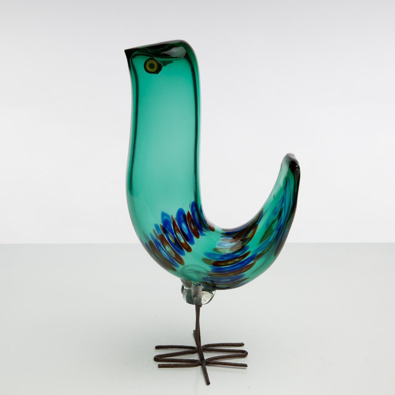 Glass bird standing on copper feet, designed by Alessandro Pianon for Vistosi Murano in 1962. 
Its body is made of green glass, embellished by blue and red murrines. Its eyes are made of hot-applied murrines. 
The object appears in the Vistosi