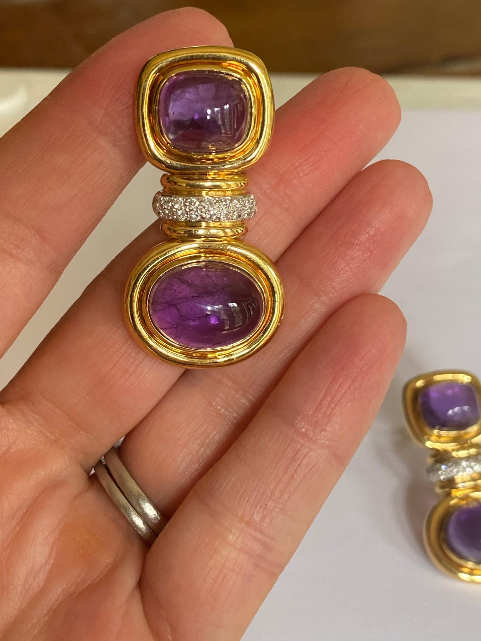 These Estate fashion earrings each feature an oval and a cushion cabochon purple amethyst encased in 18kt yellow gold separated by eighteen round diamonds and secured with omega backs. The earrings measure 1.5 inches long and weigh 27.1 grams. The