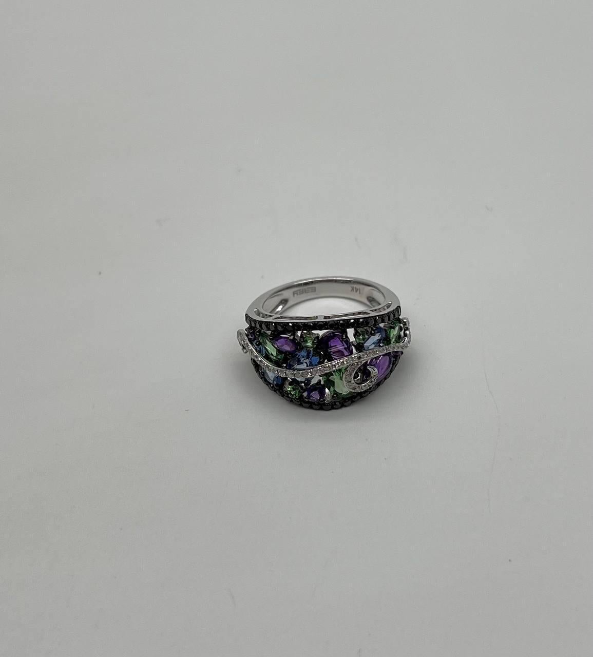 This striking ring features a bold design with an array of multicolored gemstones, including purple, green, and blue hues. The stones are set in a sweeping pattern, accented by a delicate, swirling band of smaller clear diamonds. 14K White Gold.