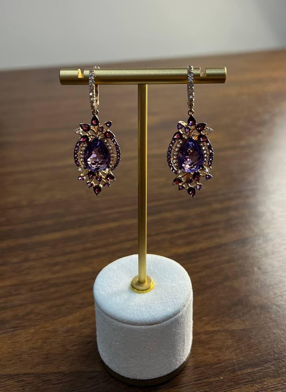 Exquisite dangle earrings featuring a captivating design. The centerpiece is a stunning purple amethyst, surrounded by intricate detailing of smaller diamonds and gemstones. The earrings are crafted with precision, showcasing a harmonious blend of