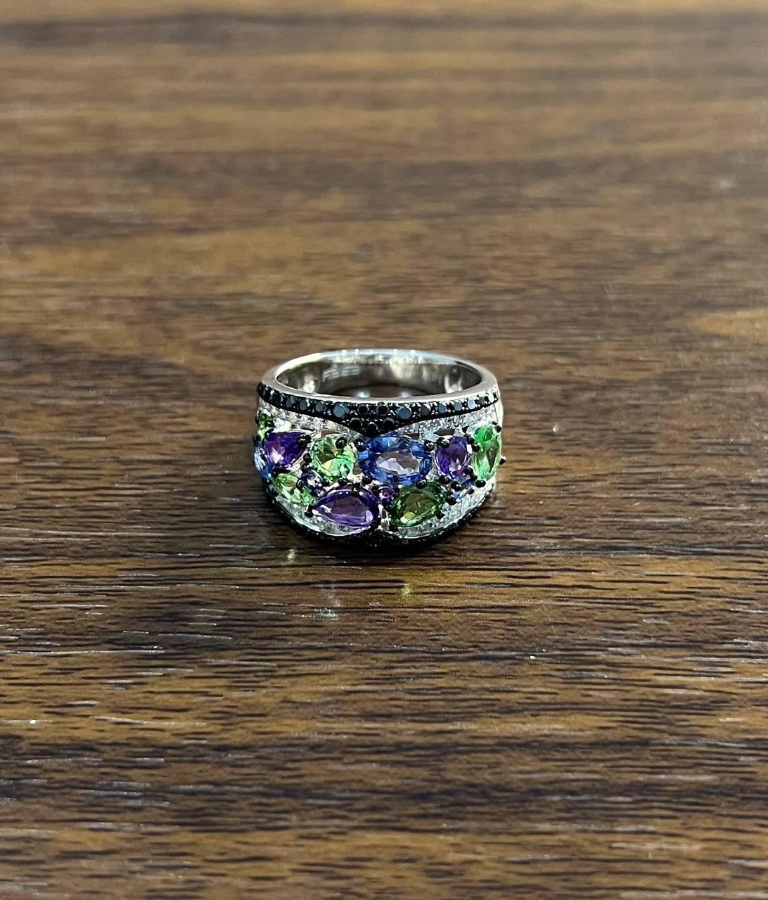 A vibrant display of color and form.
This striking ring features an array of vivid gemstones in a harmonious blend of green, purple, and blue hues. The stones are elegantly set in a sweeping design, accentuated by a band of black stones that adds