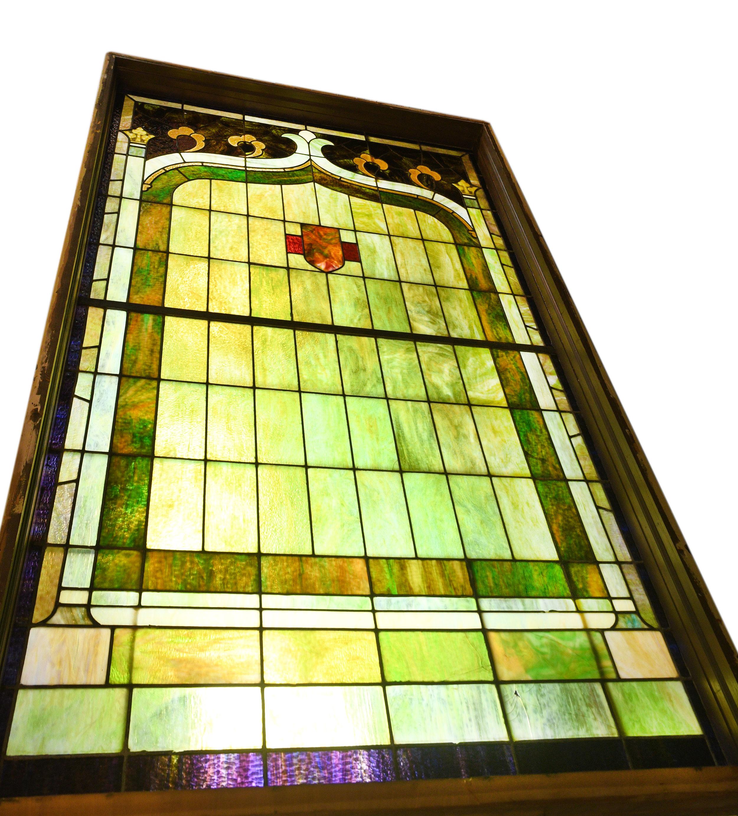 This impressively large stained glass window is made of finely crafted purple and green slag glass and features geometric patterning, floral accents, and a shield design. When illuminated, the slag glass gives off a stunningly warm glow!