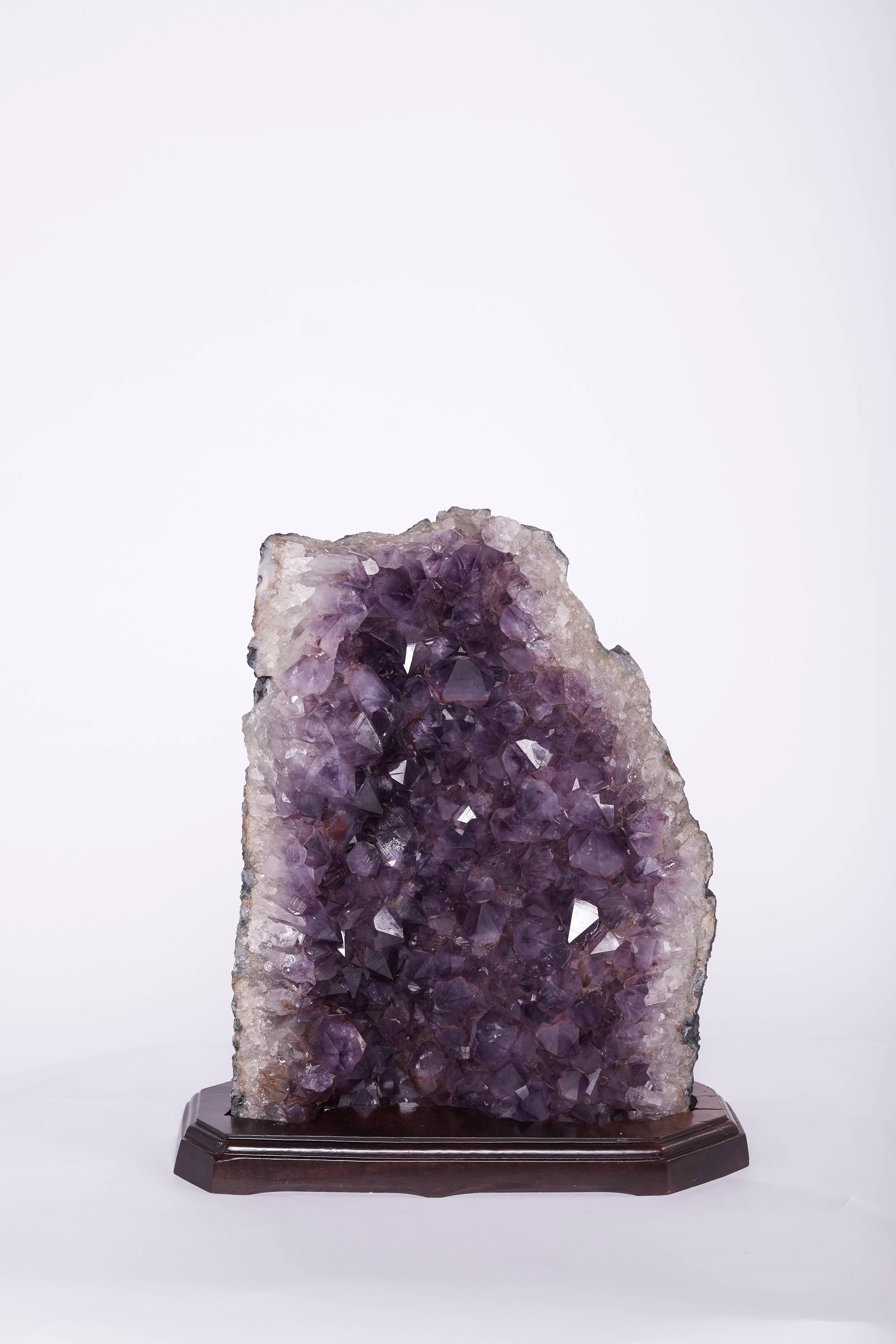 This Geode Amethyst features a free forme intense purple color and a variety of crystals. The edge of the piece is polished to reveal the white quartz layer and rests on a wood base so the amethyst is standing. Due to its intense deep purple