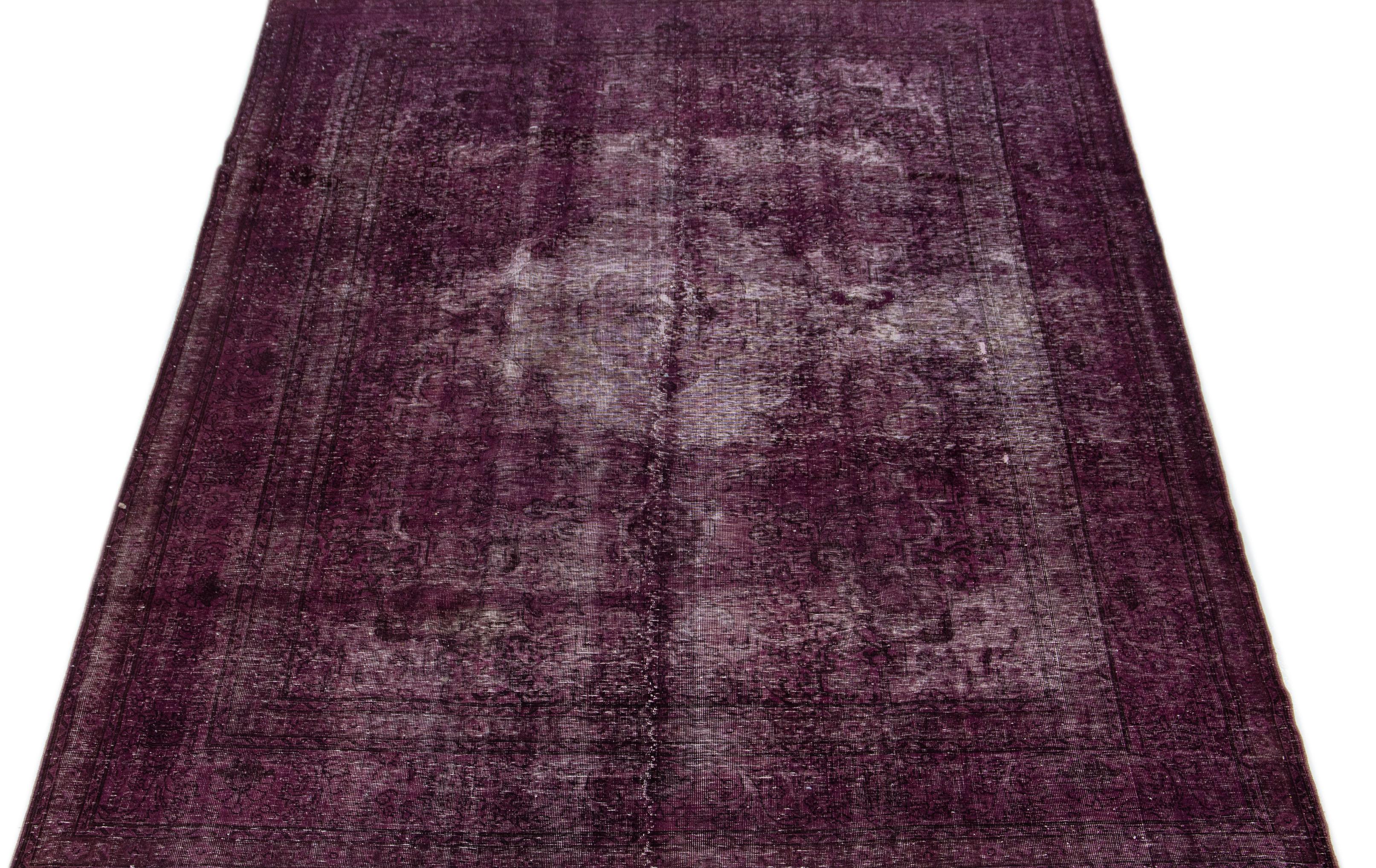 This vintage Persian rug from the 1950s showcases a striking medallion pattern in its center, which has been given an aged appearance through the process of distressing and overdrying. The rug's opulent purple color tone exudes a sense of grandeur