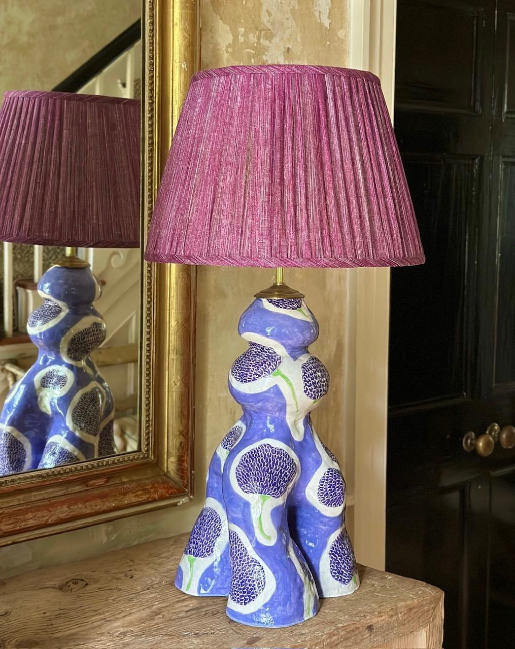 Studio-made, hand-built, curvaceous ceramic lamp form painted in two shades of purple and incised in a whimsical marigold pattern. 

Background in lilac and flowers in a deeper amethyst with spring-green stem. The form is triple-legged at the