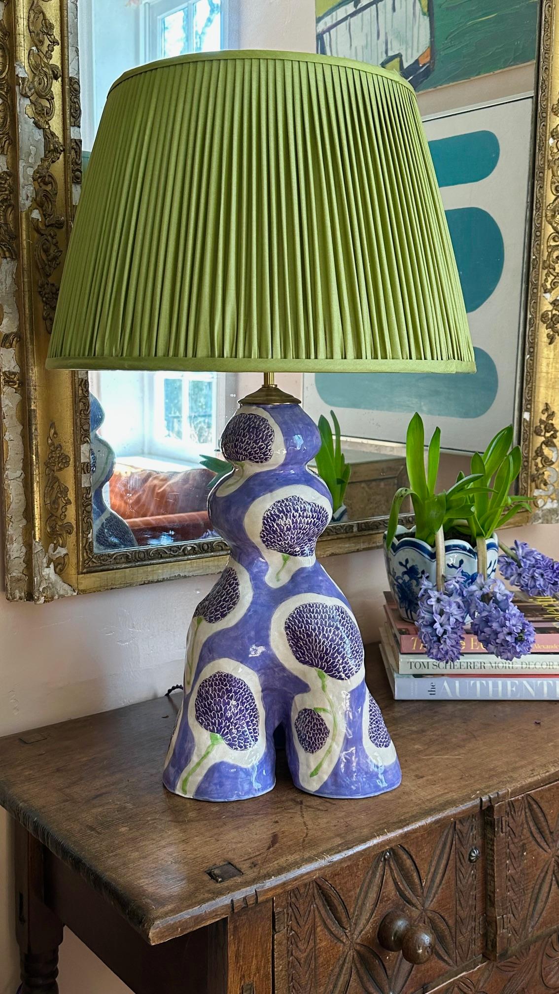 Lampe en céramique curviligne fabriquée en studio, à la main, peinte en deux tons de violet et incisée d'un motif fantaisiste de souci. 

Le fond est lilas et les fleurs sont d'un améthyste plus profond, avec une tige vert printemps. La forme est à