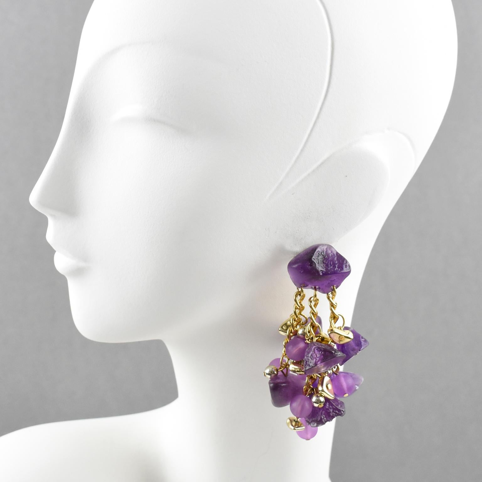 Questi splendidi orecchini a buco in lucite oversize di Studio A presentano una forma dimensionale penzolante con catena multipla in metallo dorato ornata da charms di sassolini e perline. I pezzi vantano un intenso colore viola smerigliato. Questi