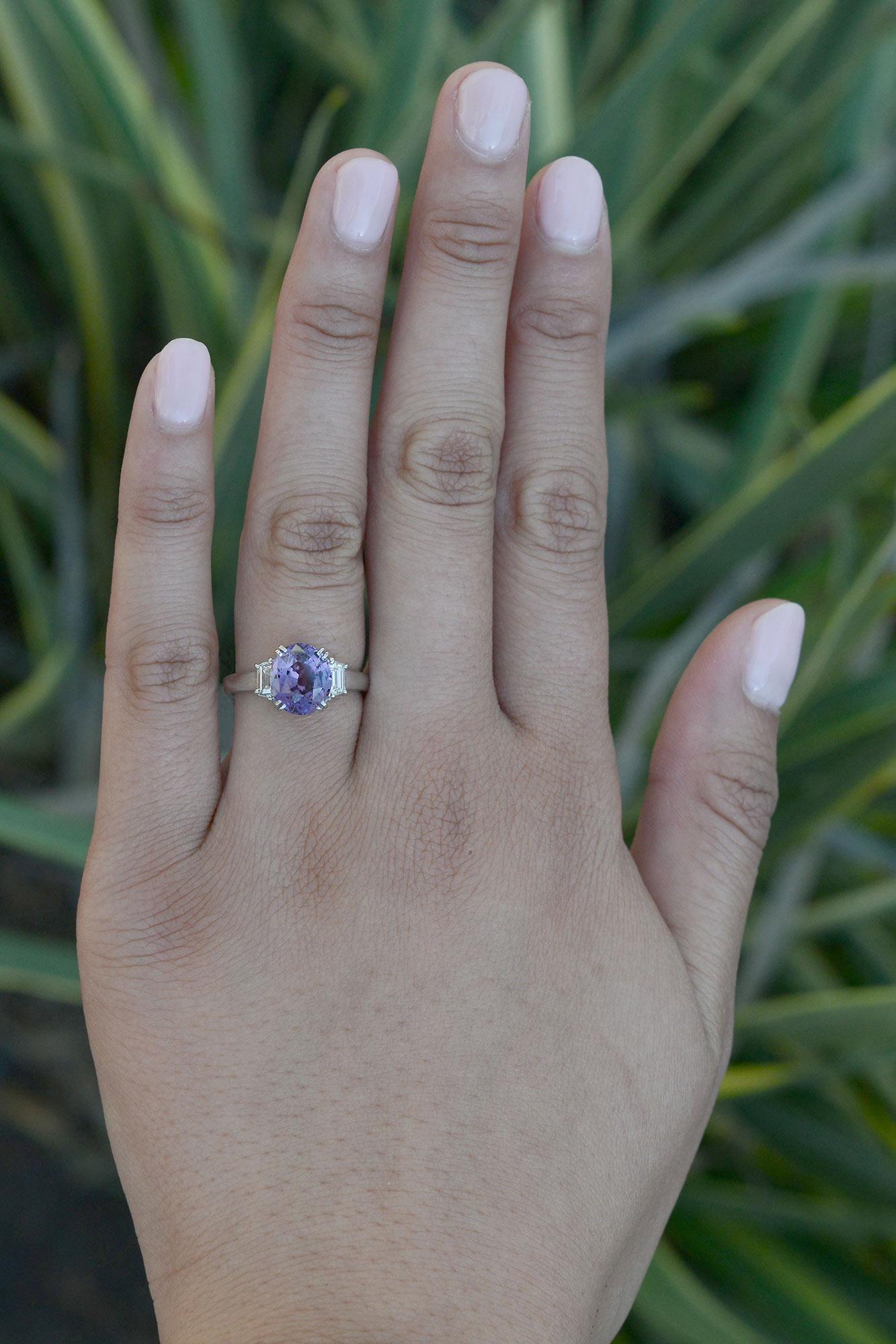 A passionately beautiful purple sapphire engagement ring in an elegant 3 stone setting. Taking center stage is a 3.23 carat captivating, brilliant and lively natural sapphire of a shimmering shade of pinkish lavender with a fabulous luster. The