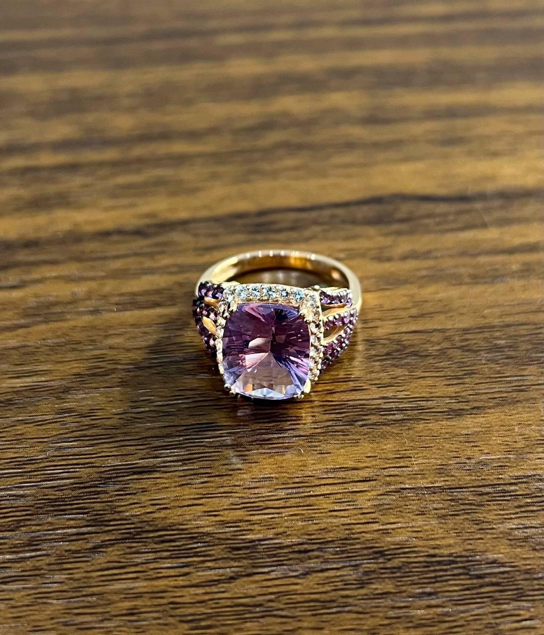 Descubre la elegancia de este exquisito anillo. Con una cautivadora gema amatista púrpura de talla cojín, este anillo es una obra maestra del diseño. La piedra central está rodeada por un halo de brillantes diamantes que realzan su resplandor. La