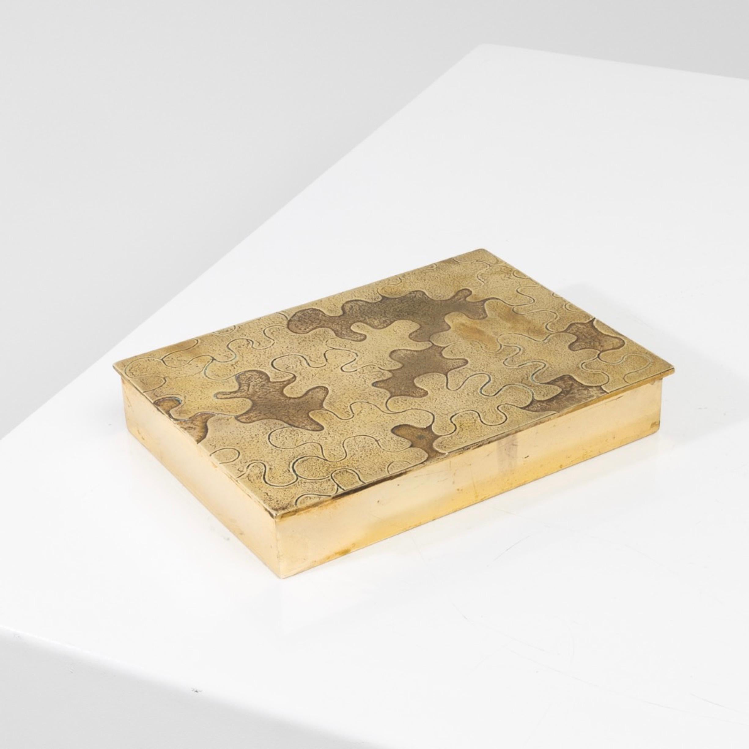Puzzle von Line Vautrin - Schachtel aus vergoldeter Bronze
Großer und breiter Kasten aus vergoldeter Bronze, dessen Deckel mit einem von Line Vautrin geschnitzten Rätsel versehen ist.
Die gemeißelten Puzzlestücke mit verschiedenen Verzierungen in