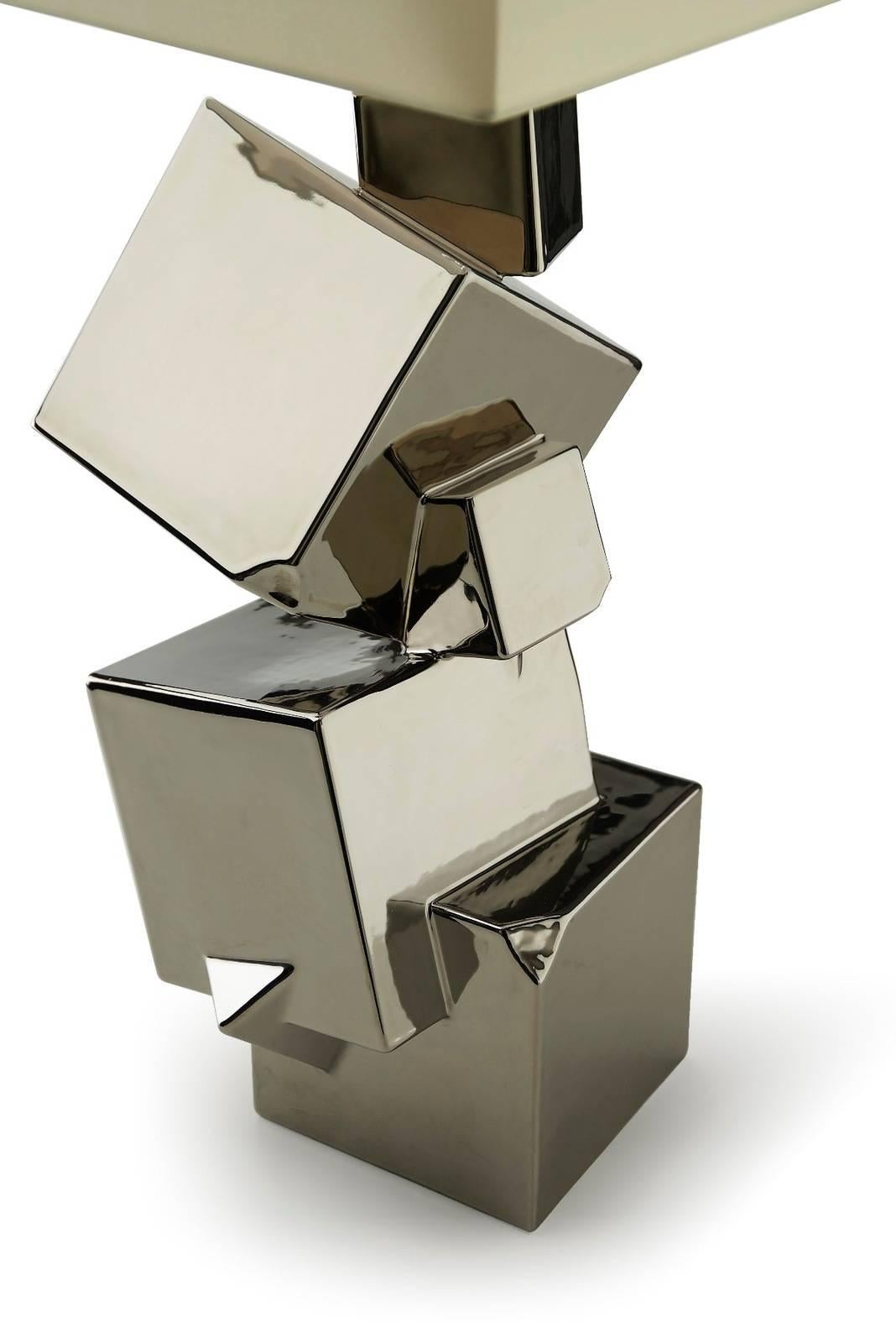 Part of the Pyrite collection, this table lamp features a rectangular shade that complements the decoration of the body made of several cubes juxtaposed at different angles. The structure is made of ceramic with a glossy silver finish and is both