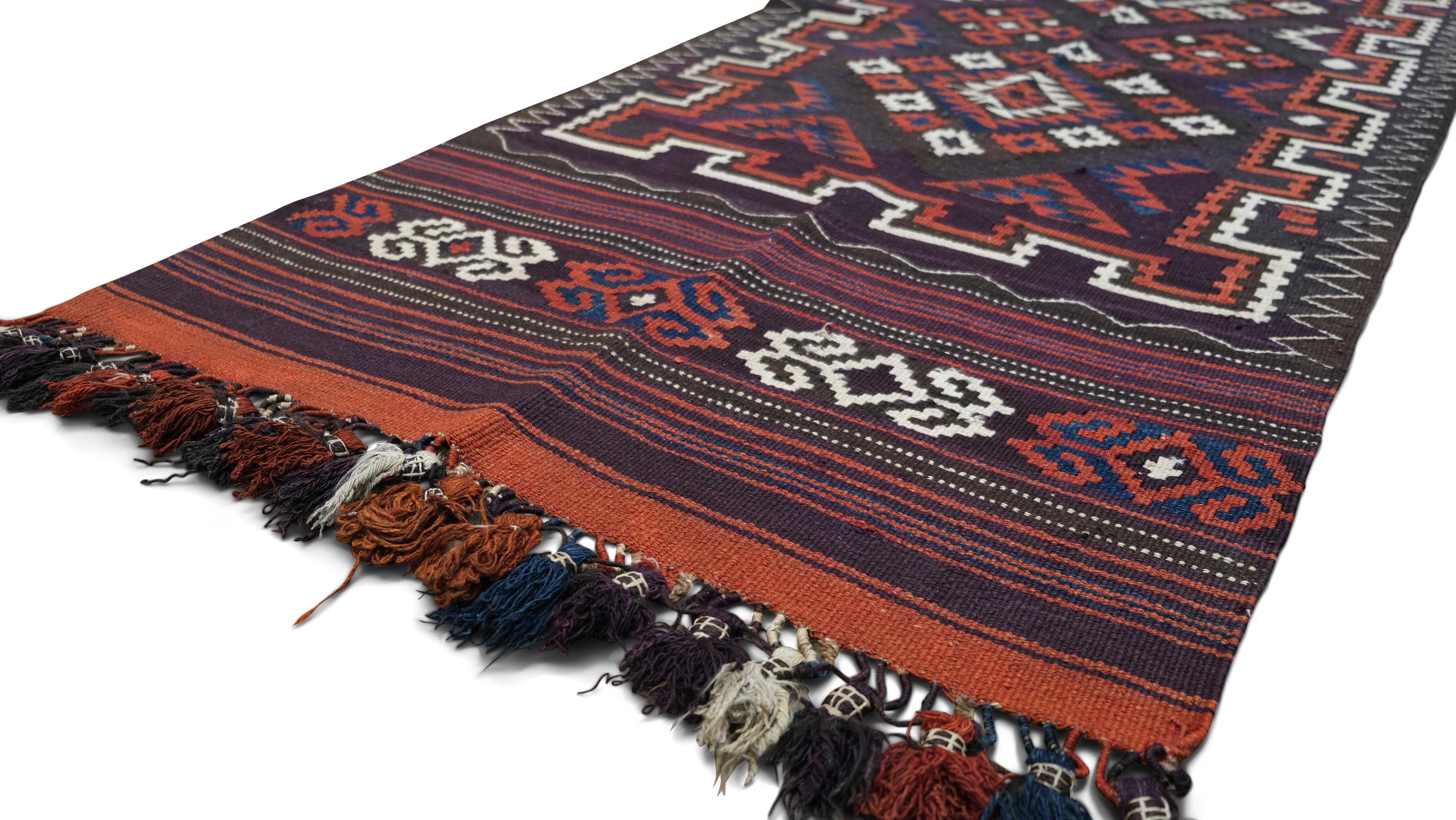 Questo runner kilim in lana tessuto a mano è un notevole esempio di tessitura tribale tradizionale, caratterizzato da un audace motivo geometrico a rombi circondato da intricati motivi e disegni simbolici. Le profonde tonalità del rosso ruggine, del