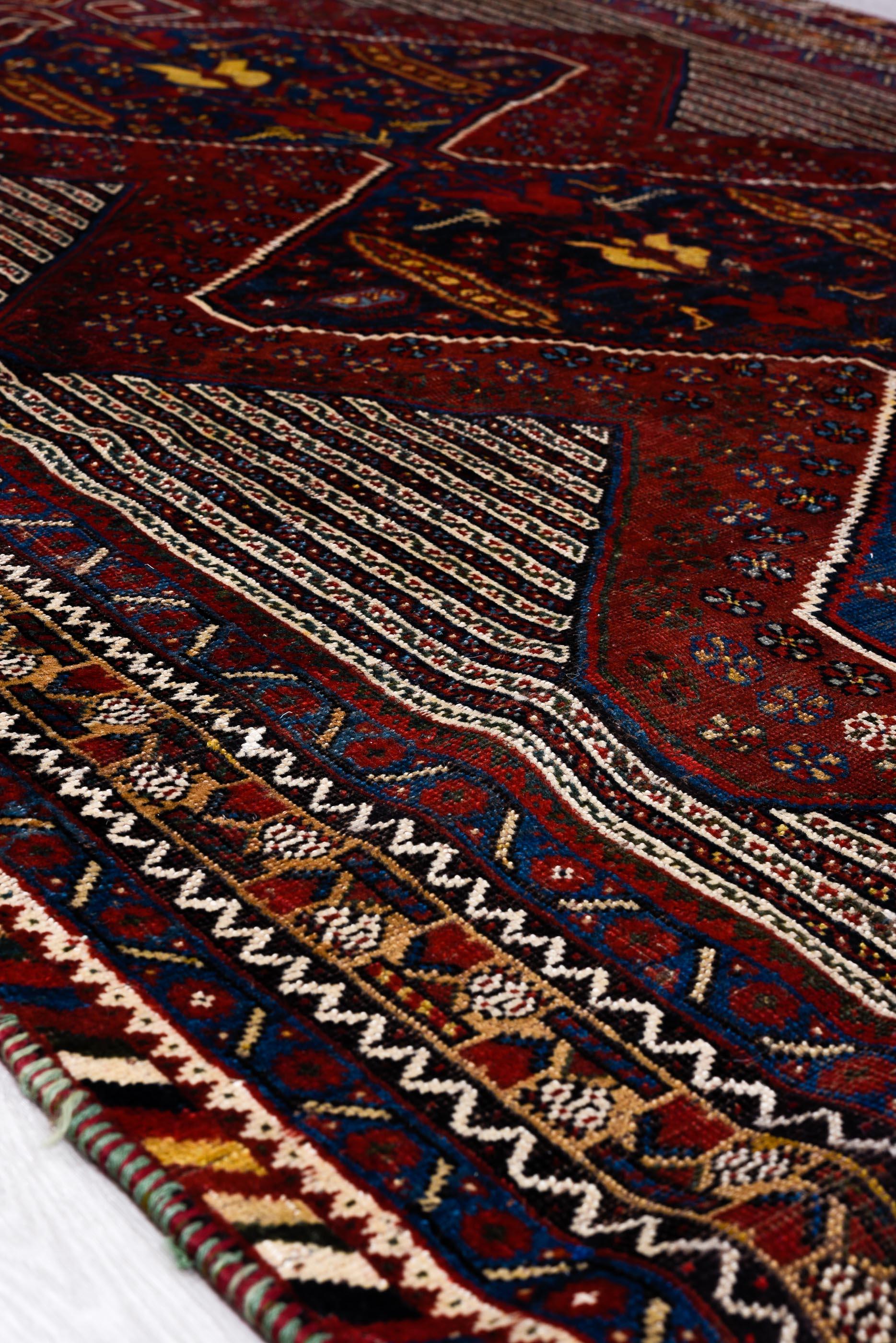 Richly composed, this Qashqai rug features three stacked hexagonal medallions in deep crimson and indigo, flanked by fine ivory ground striping. Floral and bird motifs lend a folkloric spirit, while the zigzag and serrated borders offer tribal