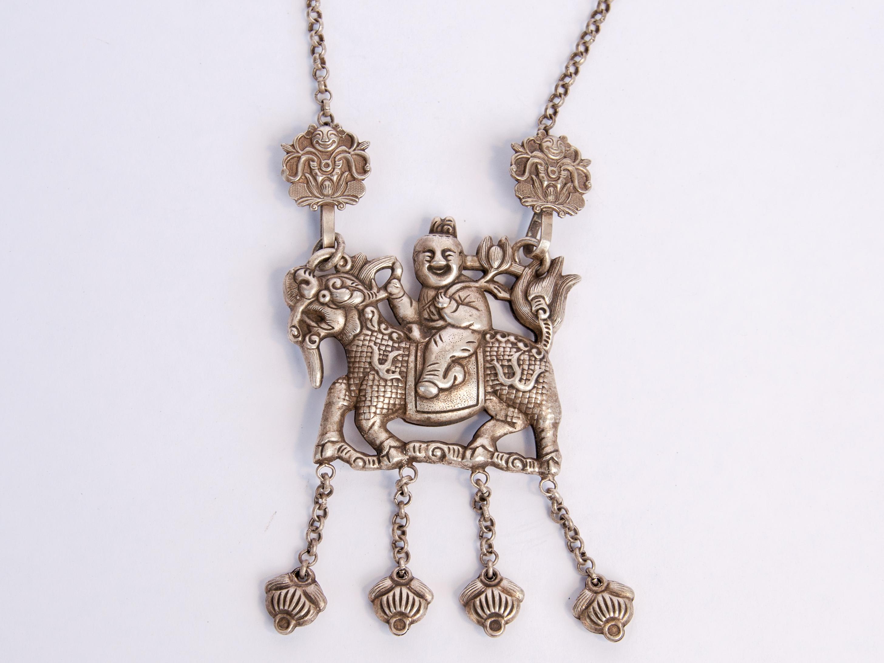 Qilin amulet necklace with infant rider, silver alloy with repousse work and hanging bells. Southwest China, early 20th century. Fastened on a chain necklace.
This lovingly worked amulet necklace depicts an infant mounted upon a Qilin. In his hand