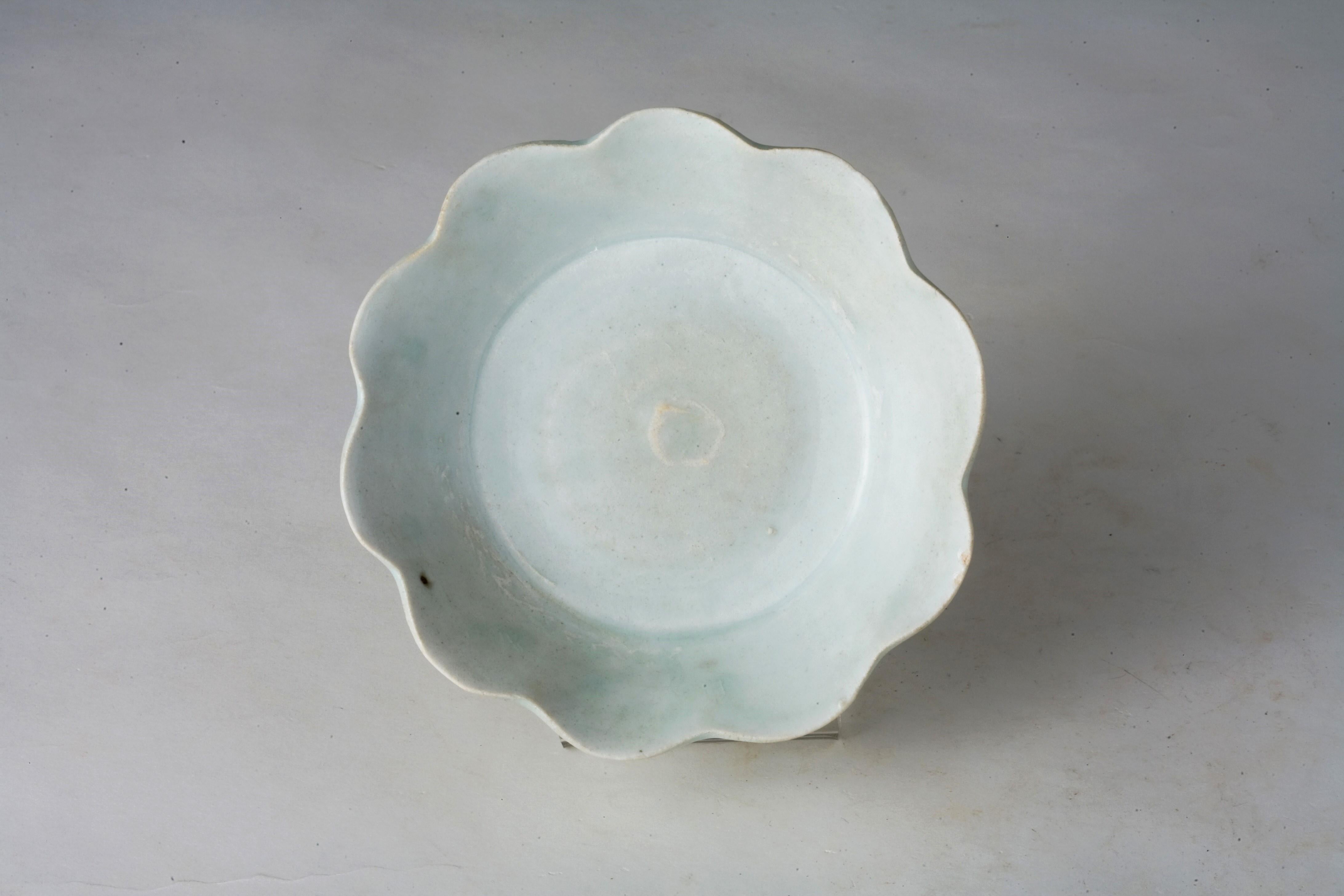 The dish has a scalloped rim, which gives it a flower-like appearance. The wavy edges are characteristic of the Song dynasty ceramics, often inspired by natural forms such as flowers. It has a pale, almost translucent blue-green glaze, typical of