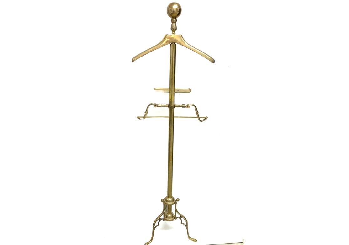 Finely constructed in brass with a cylindrical support and a large ball finial for hats over the hanger. A small shelf and a slacks hanger are below. Raised on a tripod base with ball finials on the barely twist legs ending in flat feet. The valet