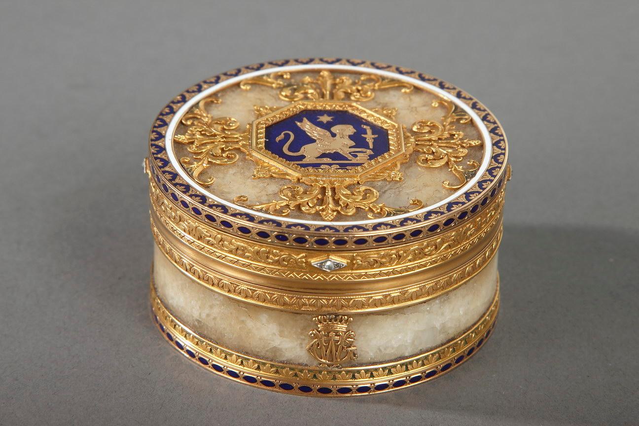 Circular quartz snuff box set in a gold and enamel mounting. The hinged lid is embellished with an octagonal medallion featuring a sphinx lying under a star in a royal blue background. The medallion is framed with four rinceaux motifs. The mounting