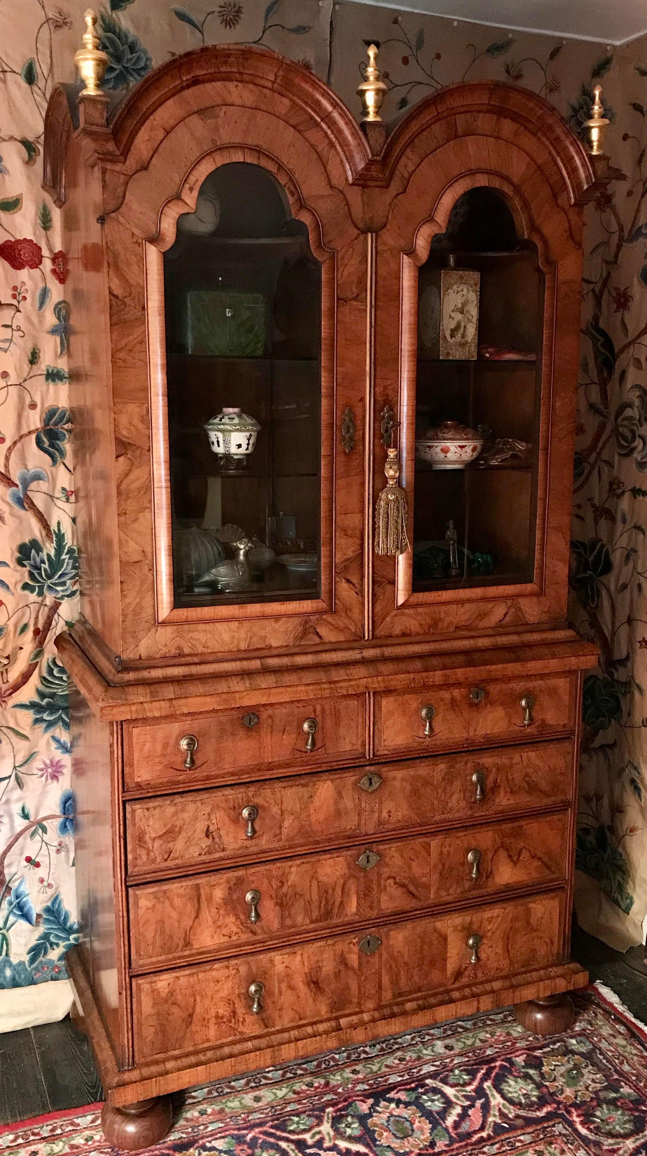 This early 18th century walnut bookcase is veneered throughout in figured walnut and is feather-banded. 

The bold cross-grain mouldings throughout are particularly well executed. The cabinet doors retain shallow, hand-bevelled glass. Bun feet