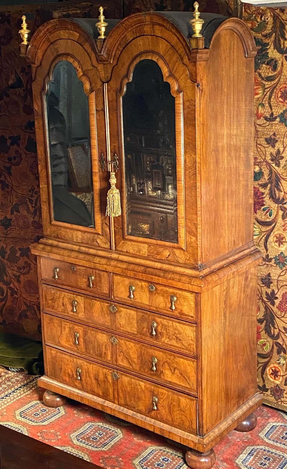A Queen Anne period walnut double-dome bookcase cabinet.

This early-18th century walnut bookcase is veneered throughout in figured walnut, and is feather banded. 
The bold cross-grain mouldings are particularly well executed. The cabinet doors with