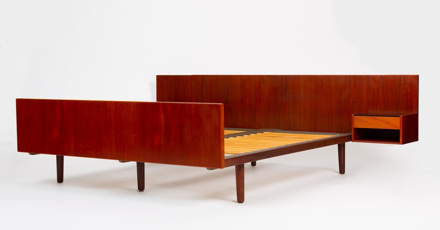A 1960s platform bed by Hans Wegner for GETAMA. The wide frame has slab-style head and footboards, with a floating nightstand mounted on either side of the headboard. Six tapered dowel legs support the frame. The slender teak base conceals twin
