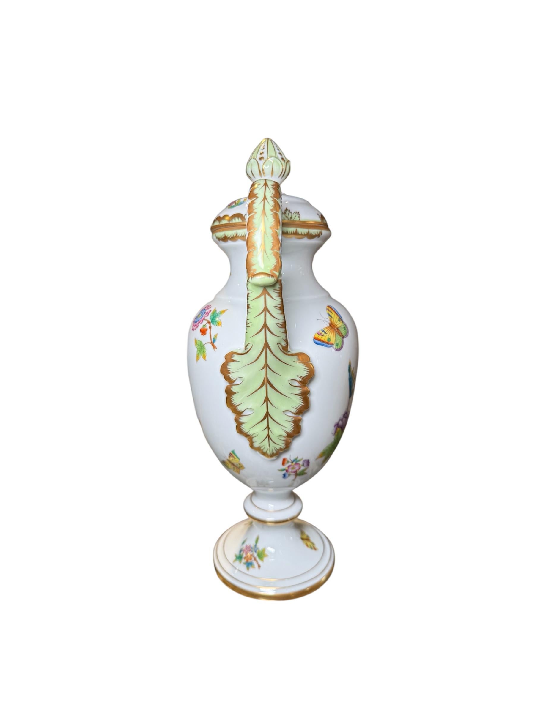 Il s'agit d'une urne en porcelaine Herend à motif de la Reine Victoria. Elle représente une urne à couvercle en porcelaine peinte à la main, décorée de fleurs et de papillons colorés, avec une bordure festonnée verte dorée. Les poignées sont