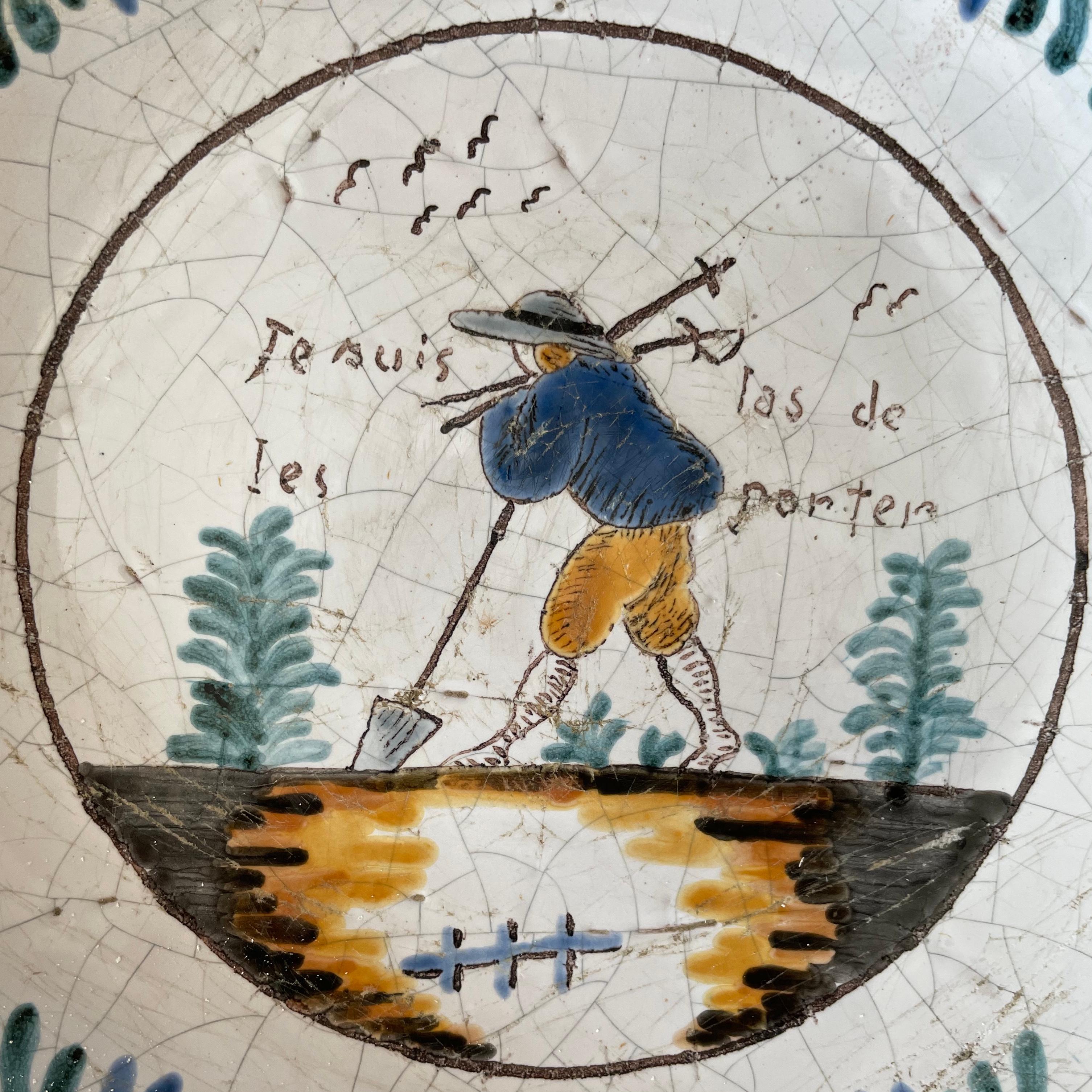 Quimper French blue and white plate. Early 19th century faience / terracotta/pottery hand painted plate with blue and yellow decoration centering on labourer in field with tools. “Je suis las de les porter,” meaning I’m done with these things. Great