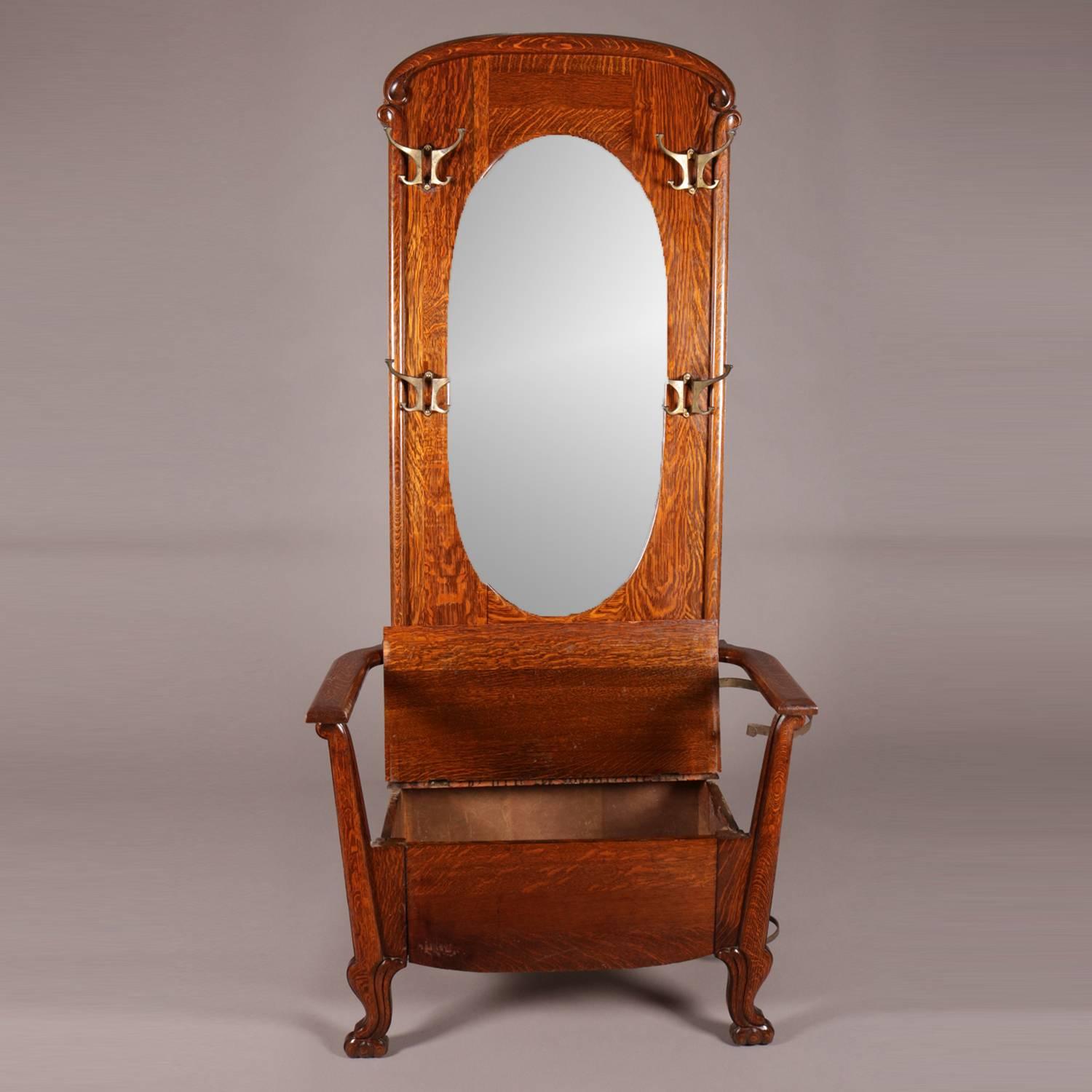 Antique R.J. Horner School carved oak chair form hall tree features scroll form crest above oval beveled mirror flanked by four bronze coat hooks, bench seat is lift top with interior storage, umbrella stand attached, seated on scroll cabriole legs,