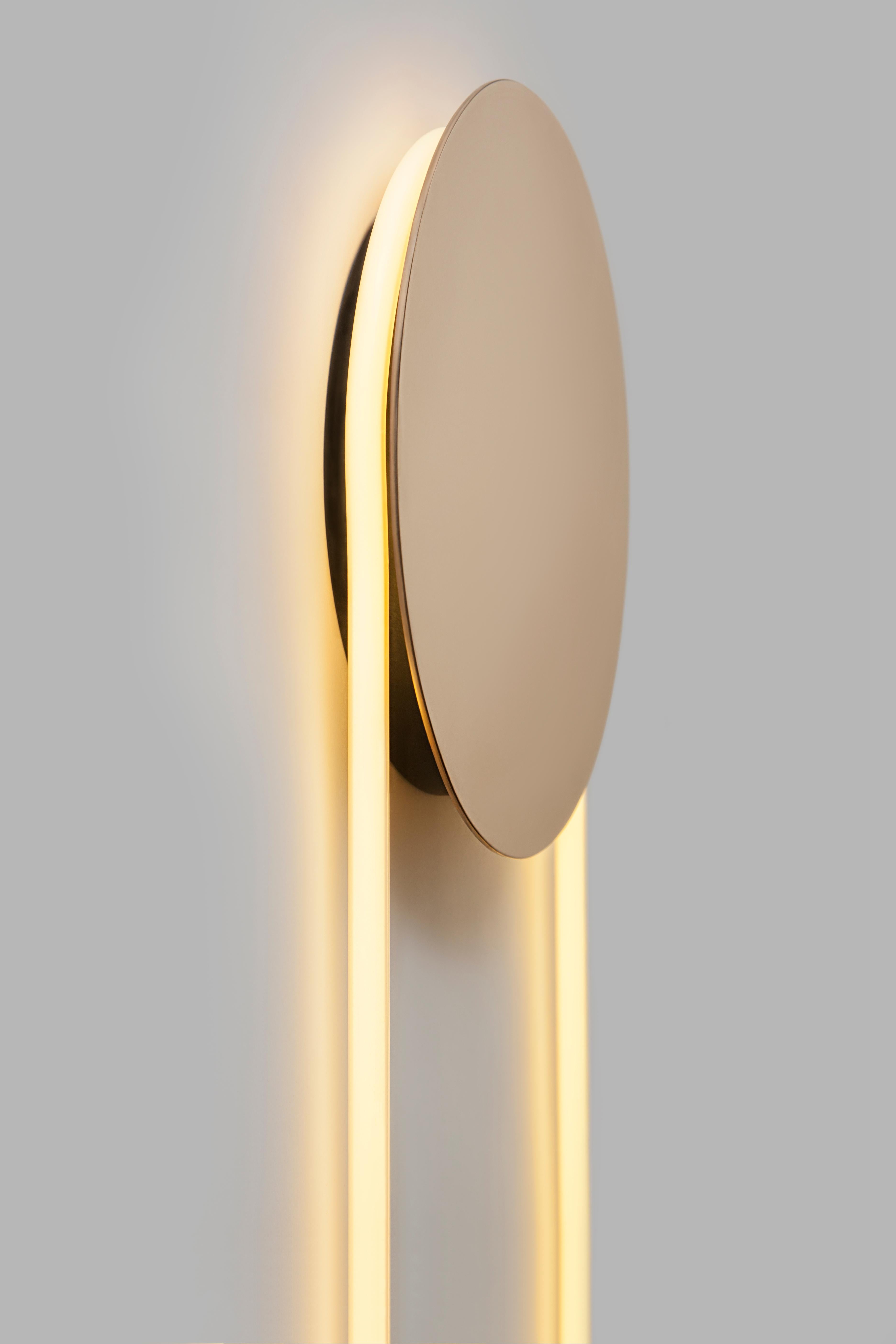 RA wall participates in its surroundings by playing with contrasts, shadows and reflections. The circular neon tube emits a bright light that reflects softly on the wall. A solid bronze disc eclipses a part of the light and draws in the hues of the