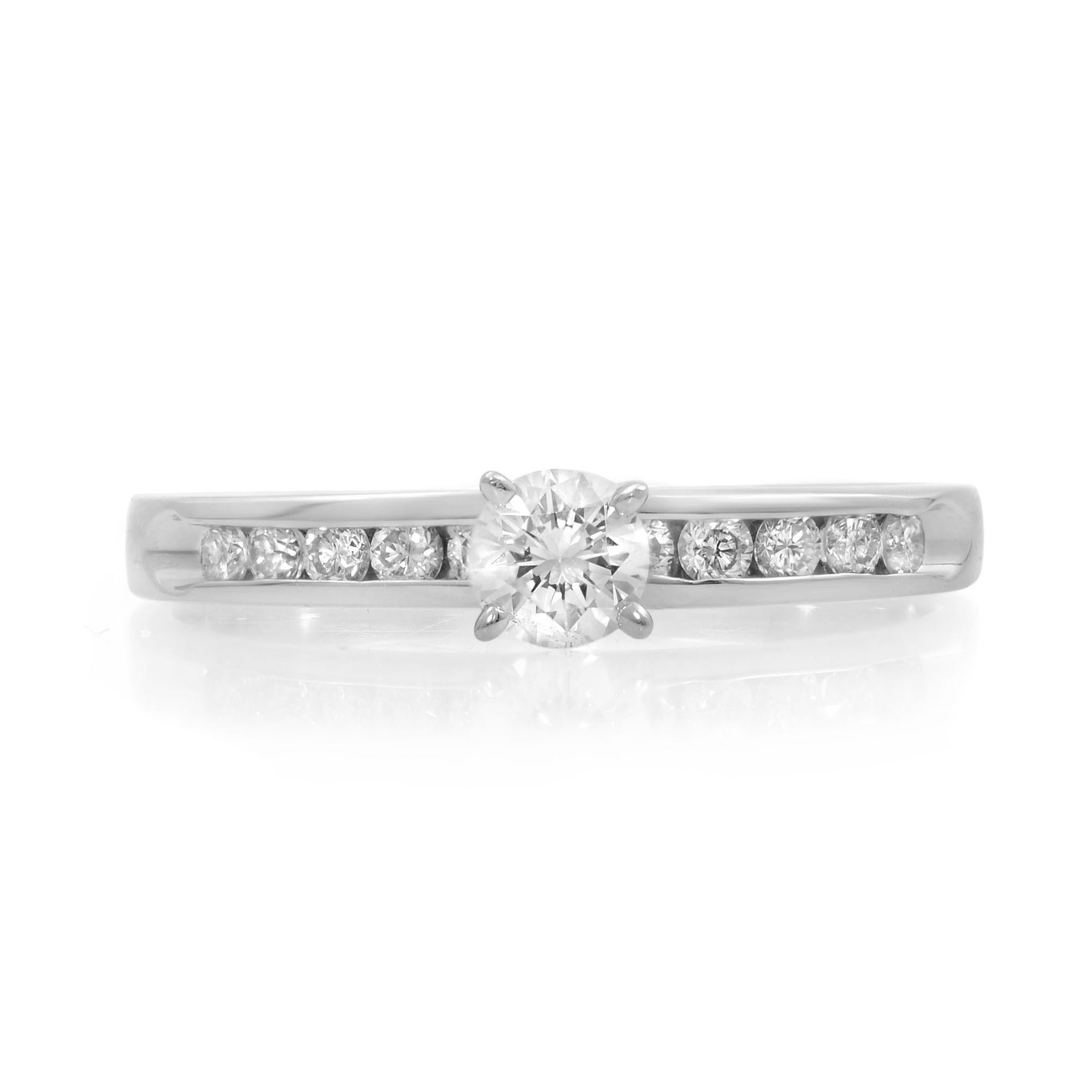 This simple engagement ring features a round cut 0.20 carat diamond solitaire, showing brilliance and sophistication. Its radiant platinum shank is embellished with channel-set diamond accents to add more beauty. It is crafted in platinum. Total