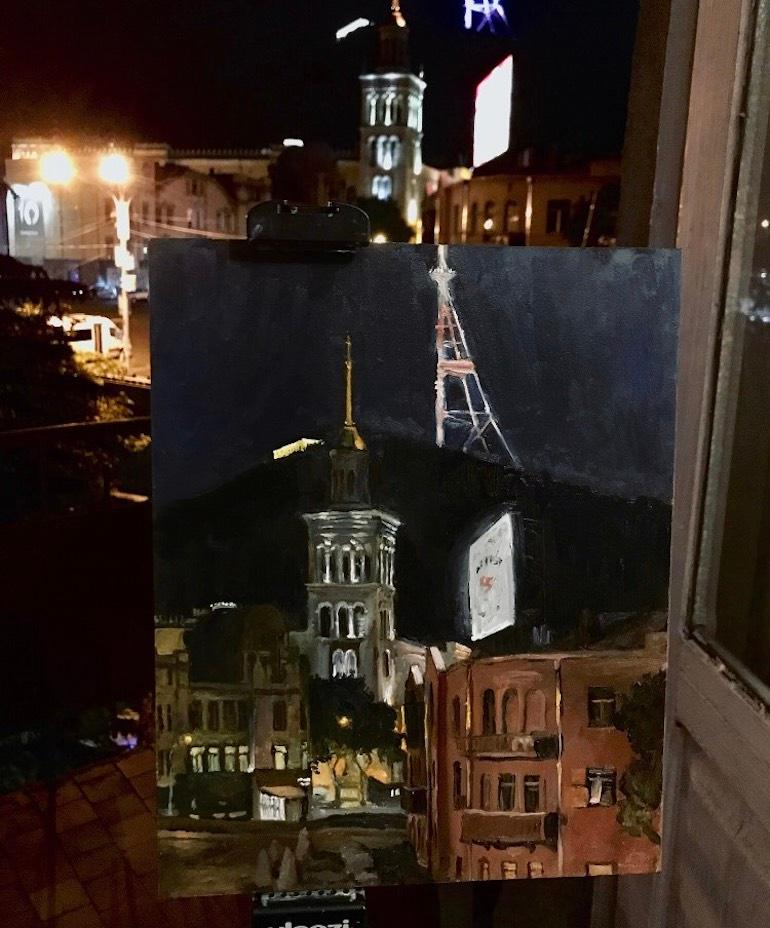 Painted from life, the artist standing on a street corner looking across to three buildings lit by ambient street lights. Rising up behind the city is a dark mass, a hill silhouetted against the comparatively brighter blue night sky.

Signed