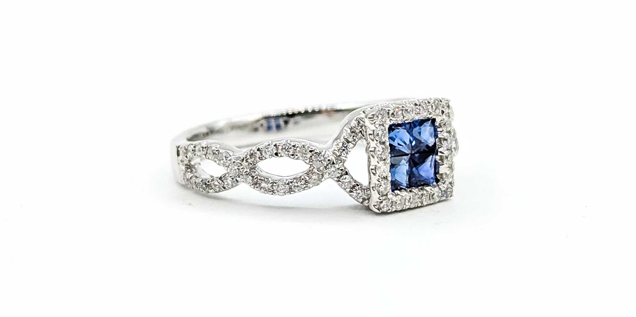Dieser elegante Ring ist aus 14 Karat Weißgold gefertigt und verfügt über einen blauen Saphir im Prinzessinnenschliff mit einem Gewicht von 0,40 ct in einer unsichtbaren Fassung, die von einem Kranz funkelnder runder Diamanten mit einem