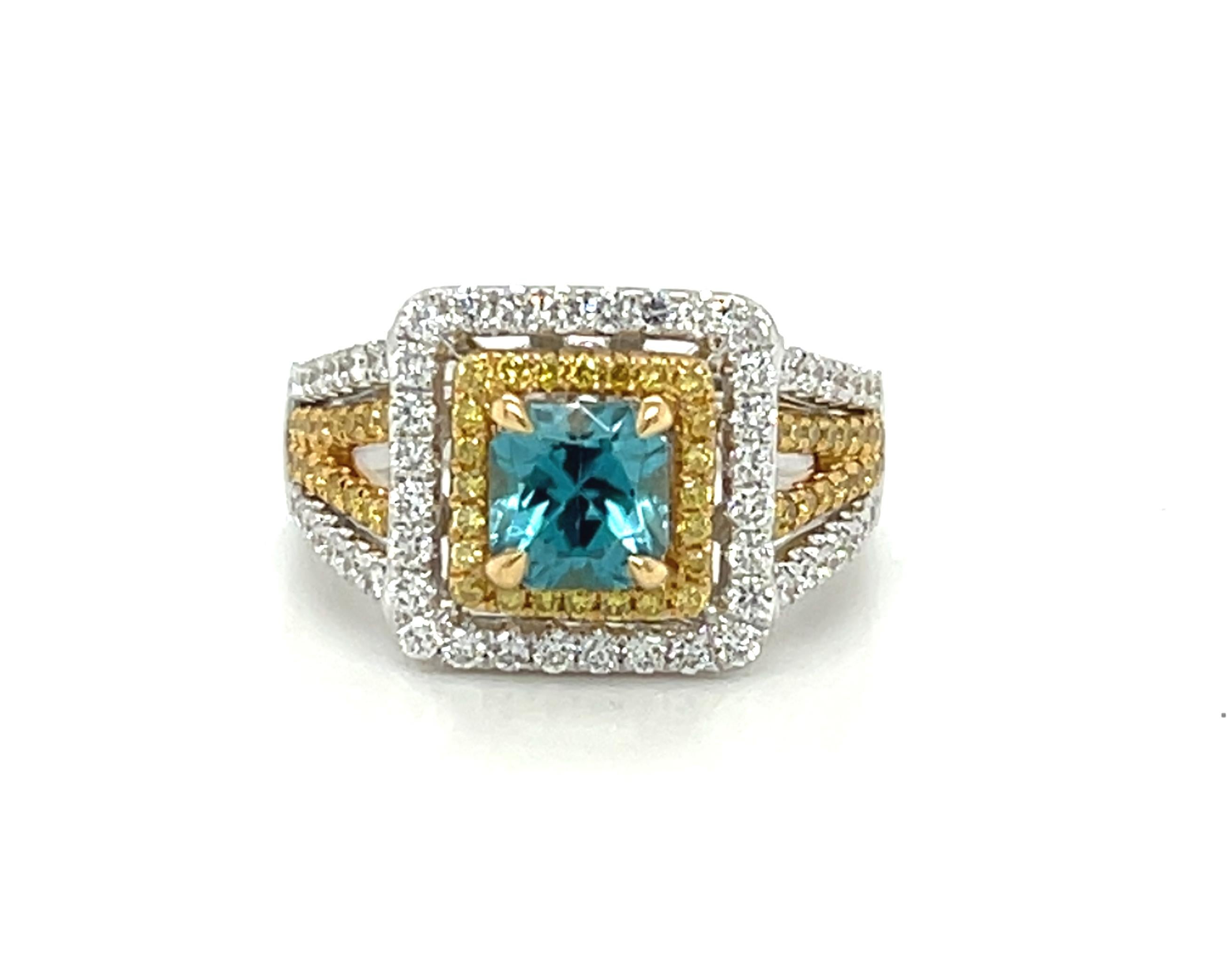 Cette magnifique bague en or jaune et blanc 18 carats présente un zircon bleu brillant encadré d'un double halo de diamants blancs et jaunes fantaisie étincelants. Le zircon bleu a une couleur bleu turquoise vive et brillante et a été taillé sur
