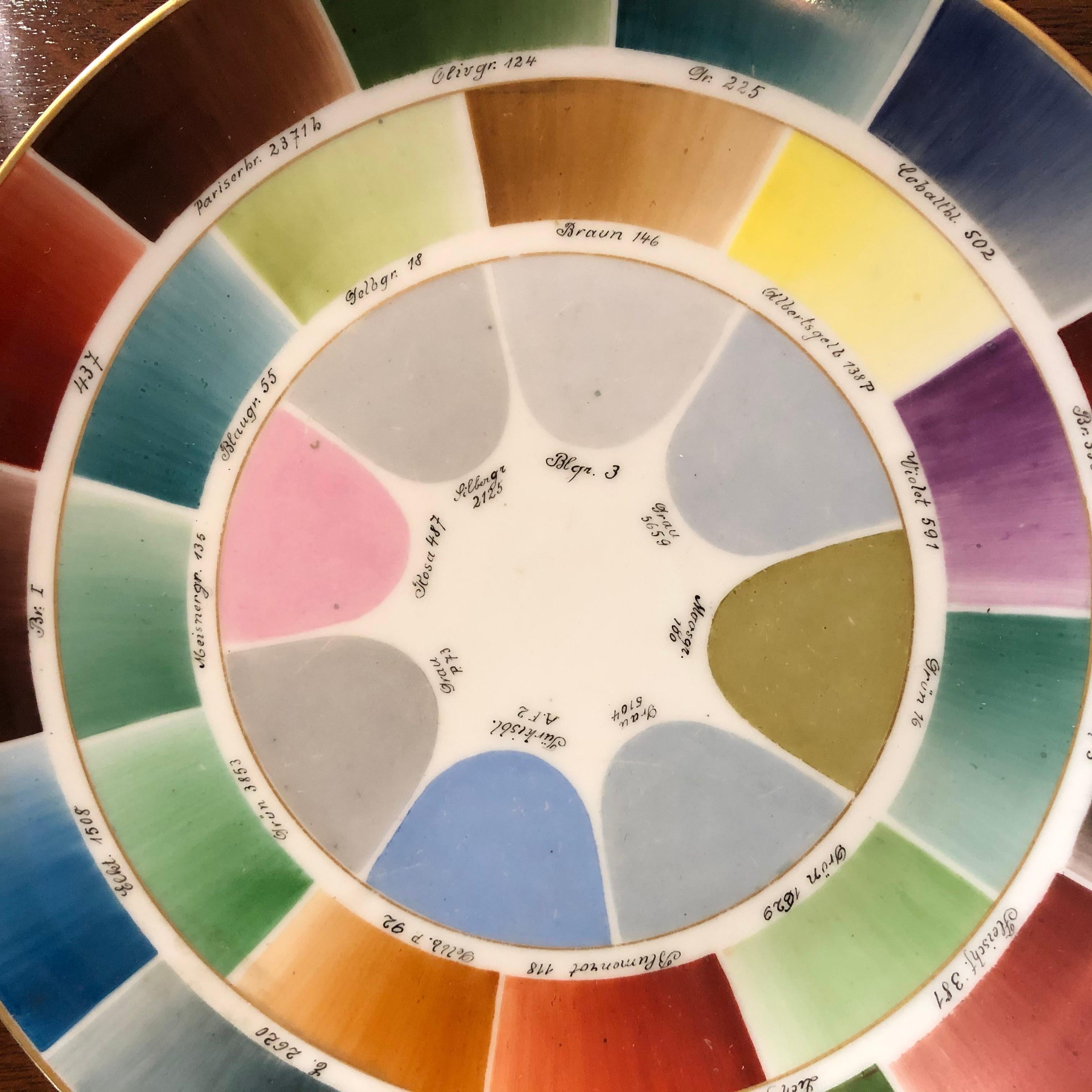 A rare porcelain factory color sample plate, hand painted with a gilded edge, showing the wide range of colors across the spectrum available, in the form of a color wheel, each one shaded and identified with its name in German. The porcelain plate