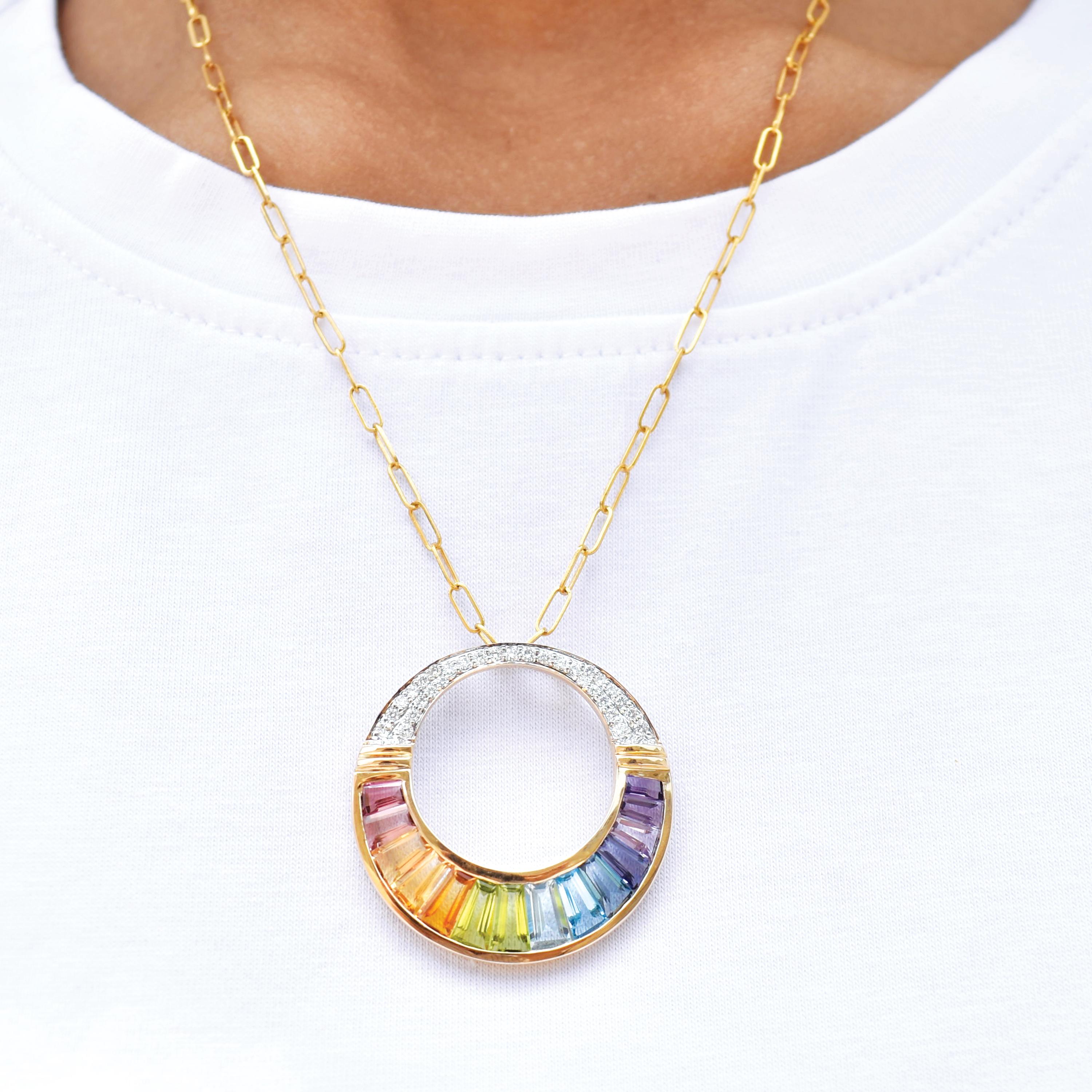 Ce pendentif circulaire en or 14 carats présente un arc-en-ciel vibrant et entièrement gradué de pierres précieuses naturelles méticuleusement serties dans un design moderne et élégant en forme de cercle ouvert. Chaque pierre de taille baguette a