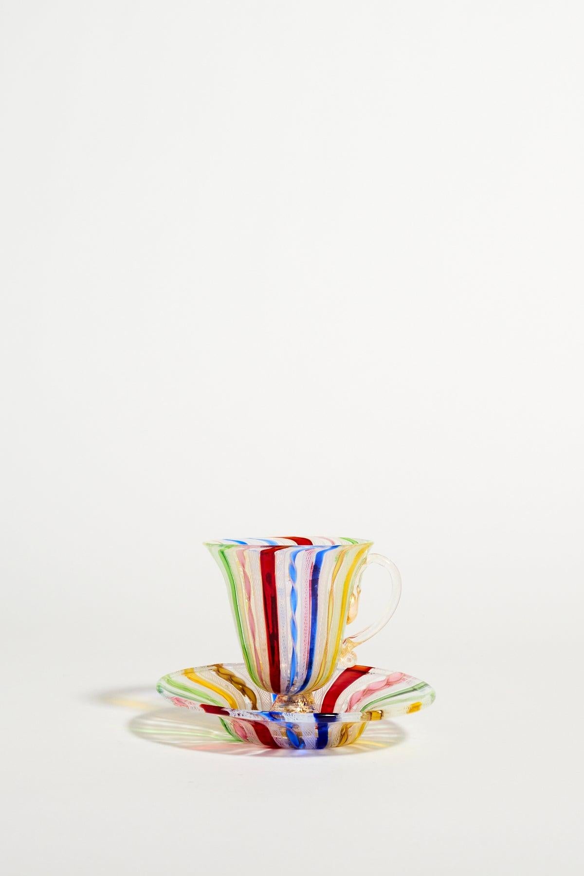 Elegant Venetian glass pedestal teacup and saucer decorated with multicoloured ribbons and white lattice and an ornate clear glass handle.
 
