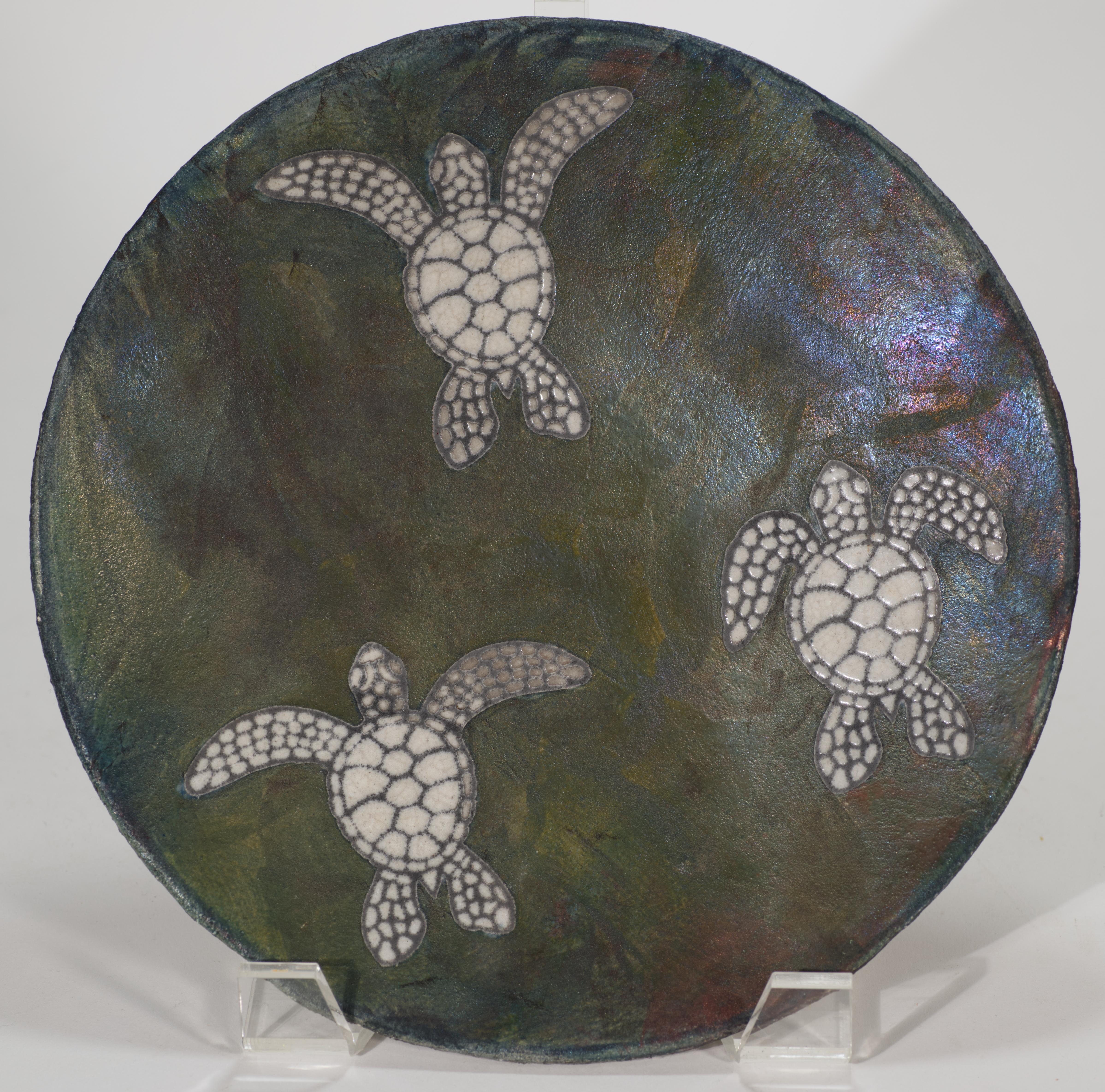 
Der dekorative Teller oder die Schale aus Raku-Keramik zeigt schwimmende Schildkröten in halbglänzender, cremefarbener Glasur. Die Glasur kontrastiert das mehrfarbige metallische Raku-Finish mit den vorherrschenden Grau- und Grüntönen und bietet