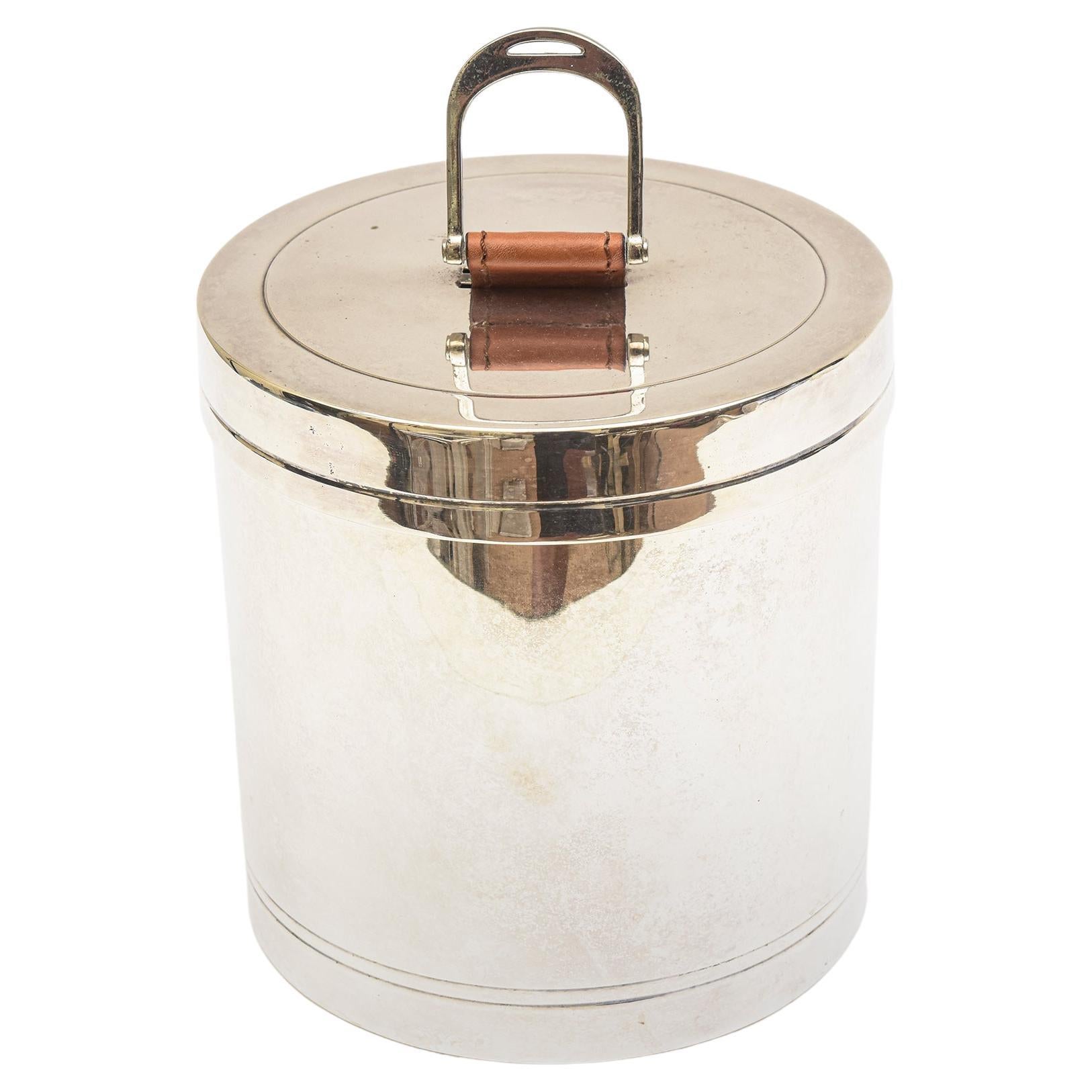This round silver plate ice bucket by Ralph Lauren from the 1990s has a combination tan leather pull handle on top with a silver plate pull. It can stand up if you push the leather down. The front of the ice bucket has a shield emblem with 3 tiny