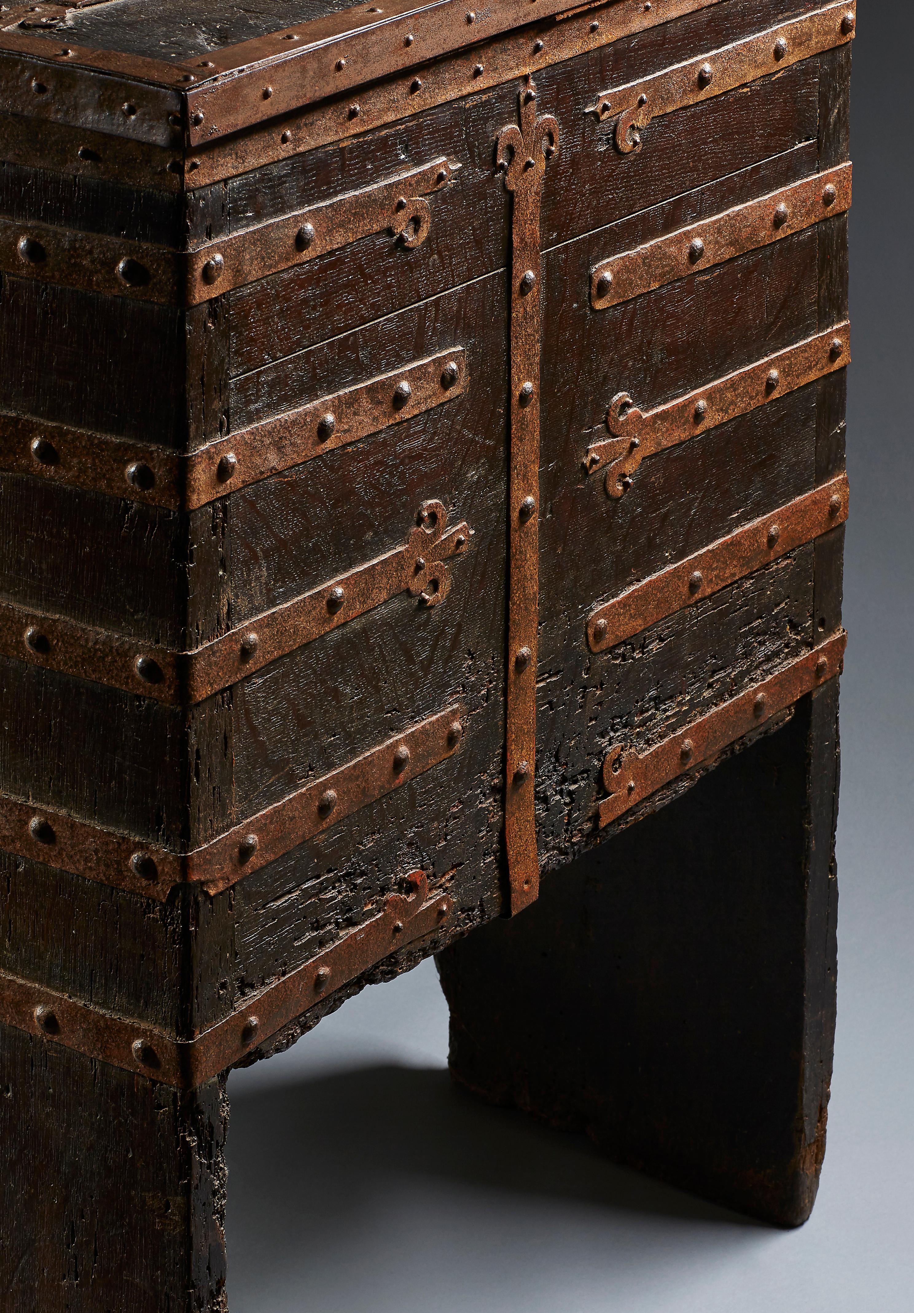 The monumental rectangular standing chest with full-height stiles, extensively mounted with wrought ironwork straps terminating in lily finials which ’wrap’ around the chest edges. These are fixed with convex head nails: running vertically up the