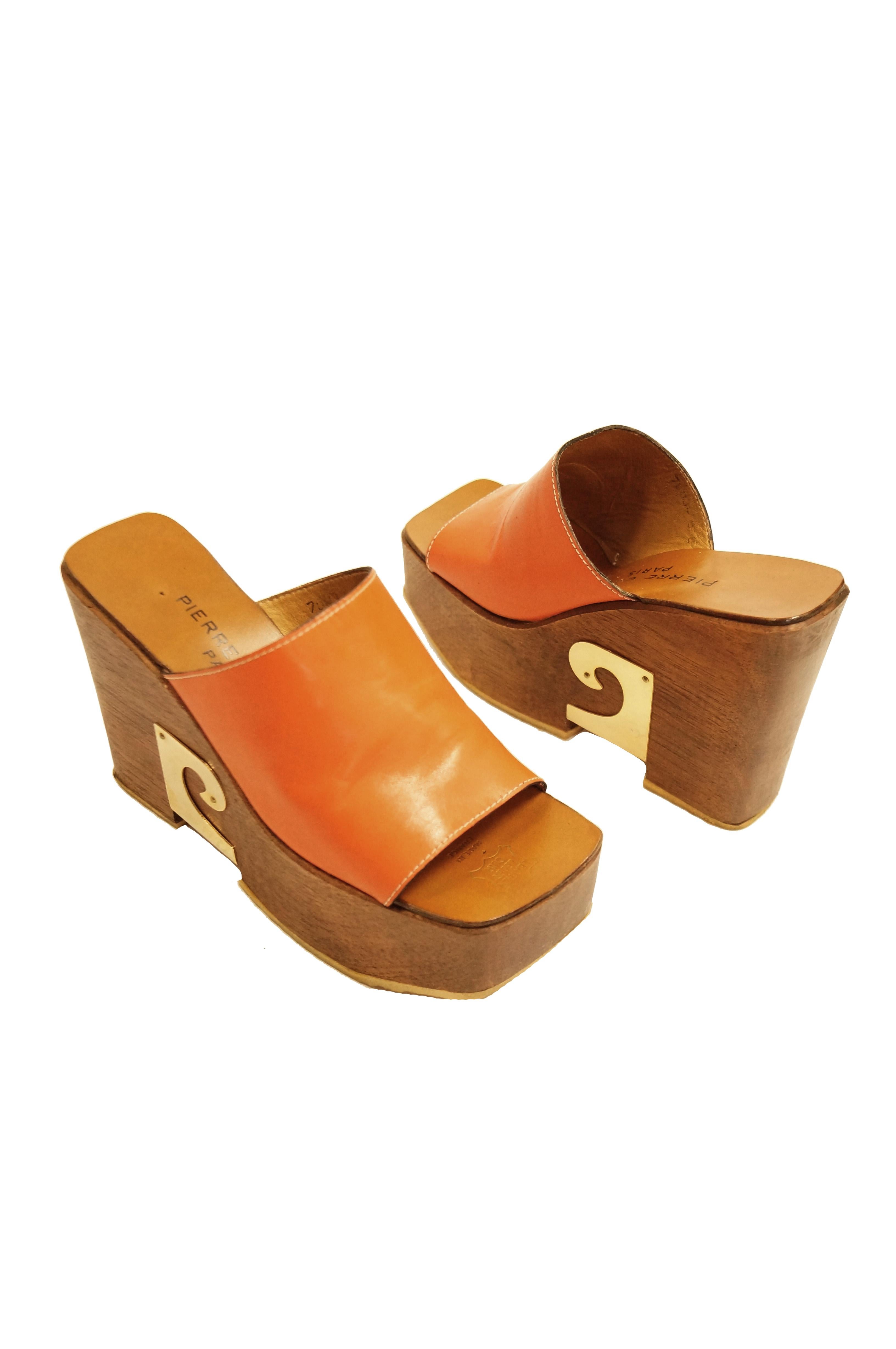 Amazing wooden clog sole shoes by Pierre Cardin. These tall platform heels feature a deep orange mule top strap and a leather sole. The thick wooden platform heels are contrasted sharply the iconic Pierre Cardin swirl logo on the outer side of the