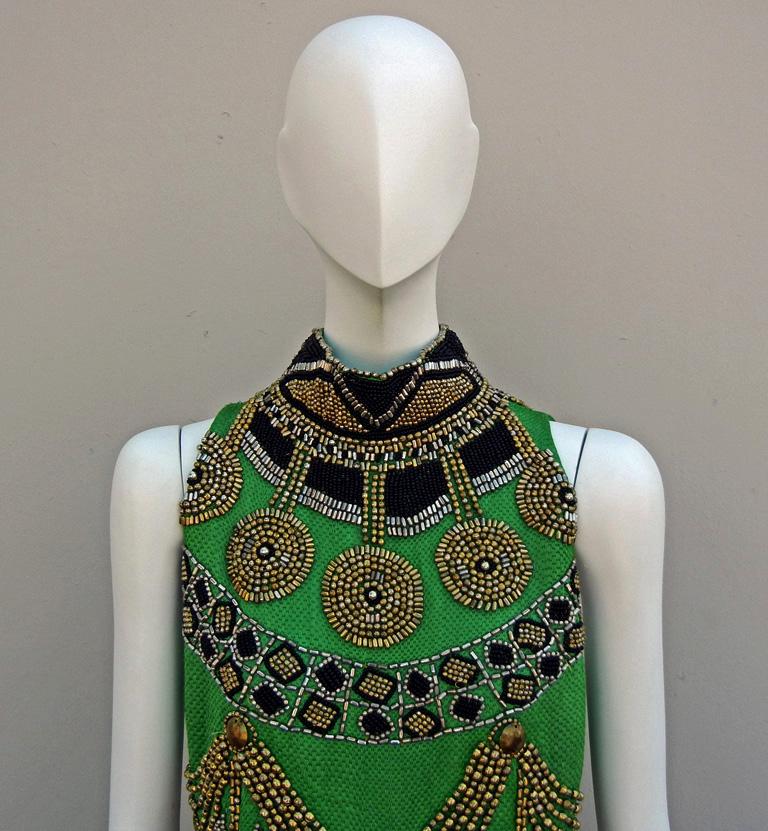 Raro abito su misura del 1990 disegnato da Gianni Versace per il suo Atelier Versace.   Abito senza maniche a collo alto in ricca seta damascata verde, ornato da pesanti perline in ottone e oro con un tocco egiziano.    Il V&A Museum ha suggerito