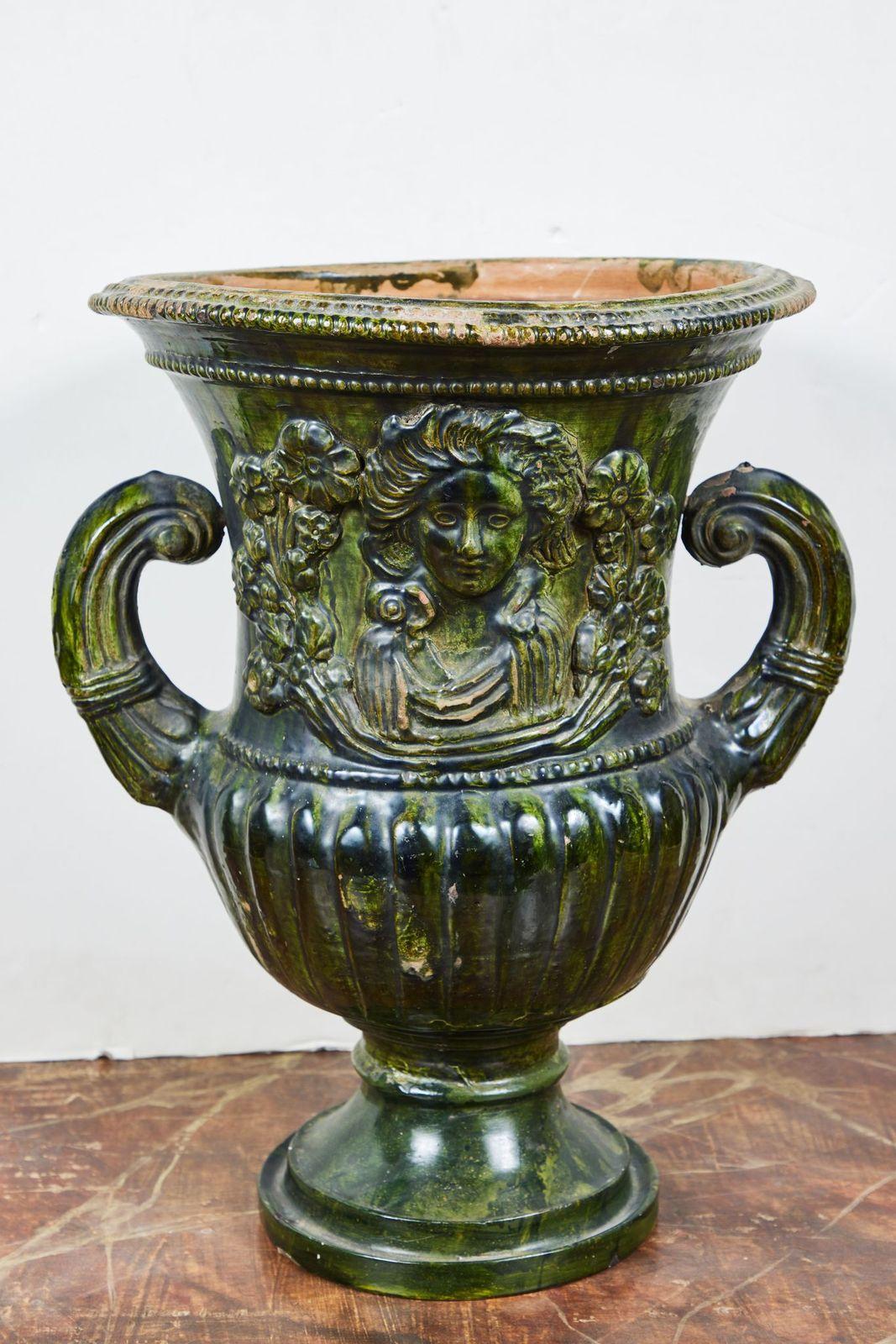 A large pair of relief molded, period, hand-sculpted terracotta urns in lush green glaze. Each piece features two, classical-style goddess-like figures in robes surrounded by foliate forms above a fluted, base tapering to a stepped foot.