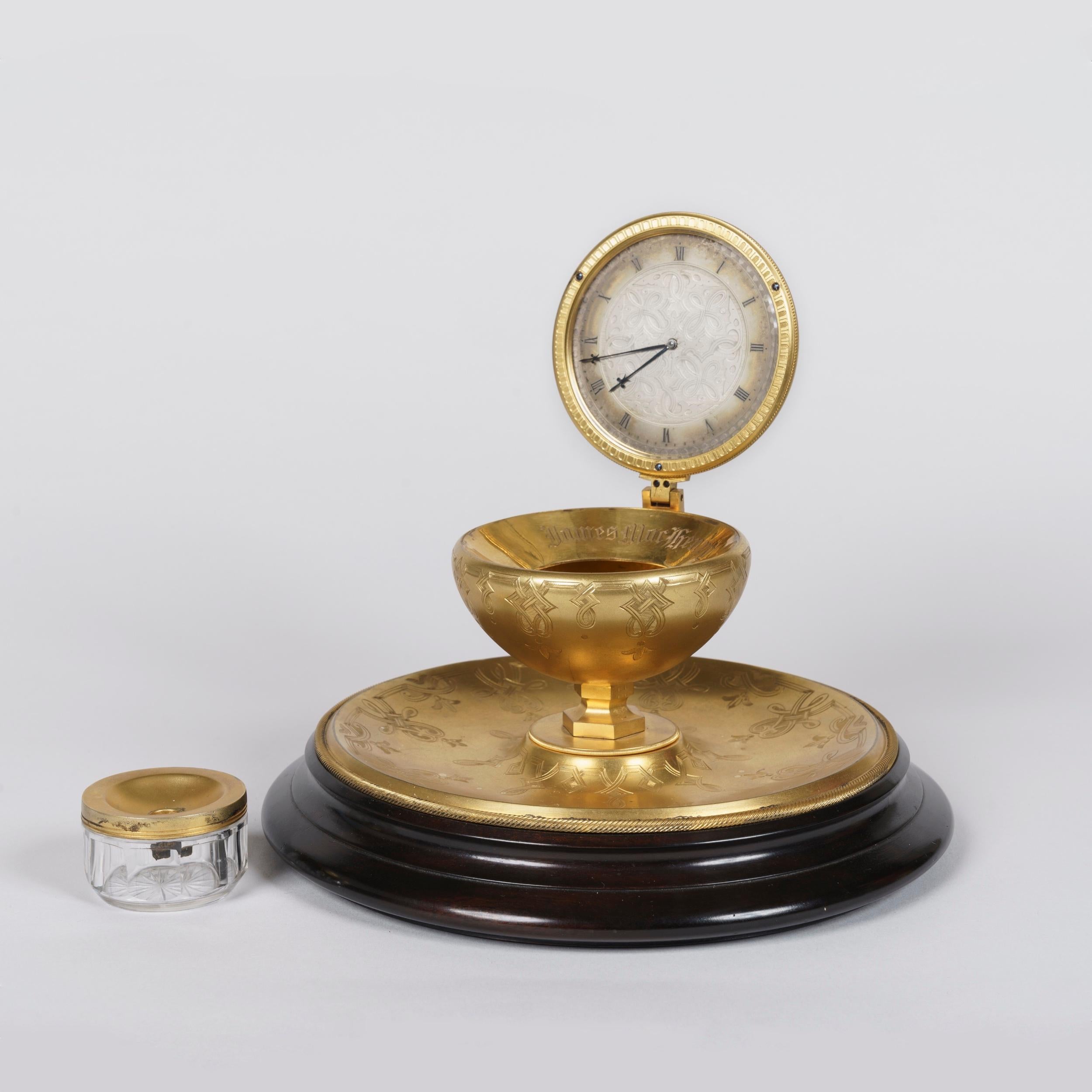 An Unusual 'Inkwell' Table Clock
Attributed to Thomas Cole

Constructed from gilt brass, the novelty timepiece rising from a turned ebonised base inset with a concave brass tray engraved with geometric strapwork; a central socle supporting the
