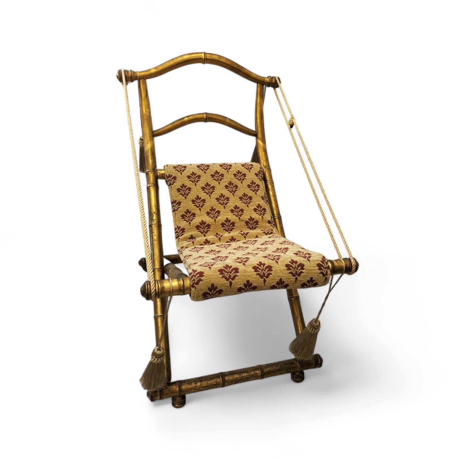 We present you with this exquisite and original 19th century folding chair, crafted from carved and gilded wood as if to imitate a bamboo form. 

This piece is elaborately adorned with trimmings and upholstered in fabric with beige, golden and red