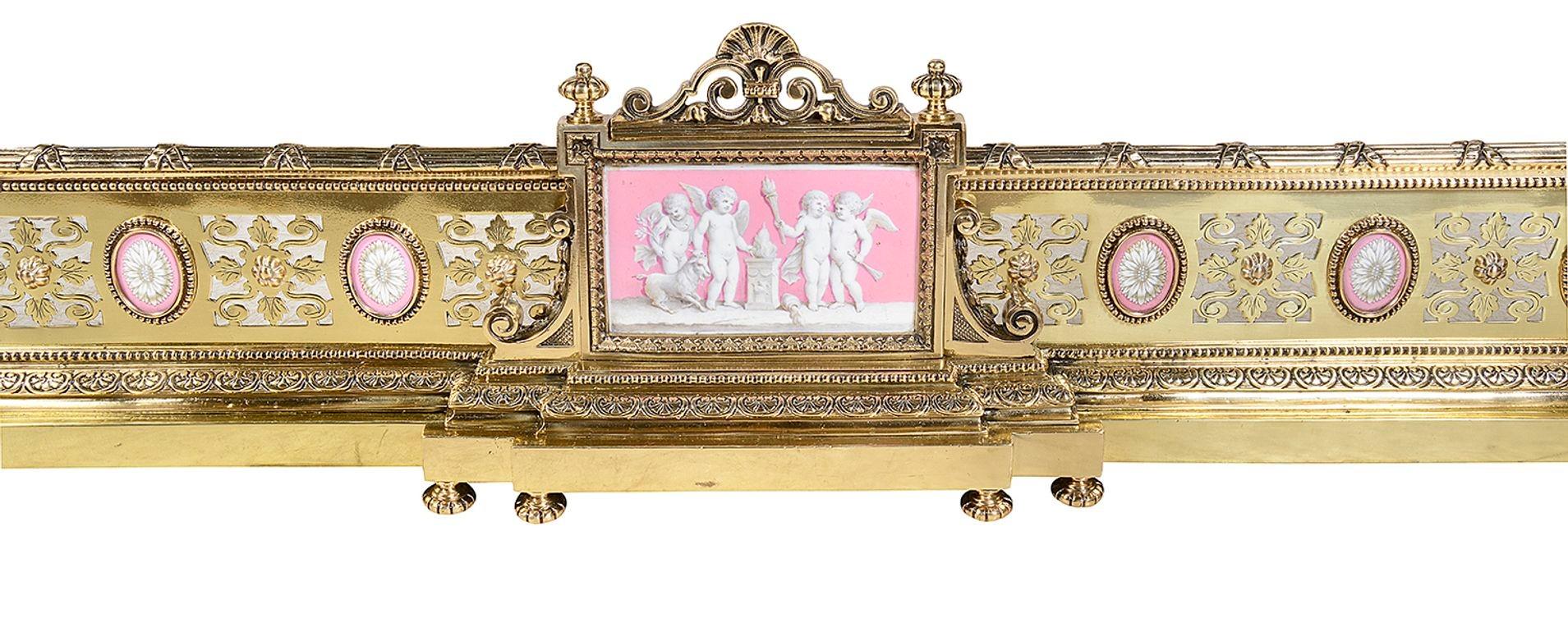 A rare fine quality French gilded ormolu fender, having classical pierced and engraved scrolling patterns, mouldings with motif and ribbon decoration. Inset pink porcelain Sevres style plaques with motifs and the central panel depicting frolicking