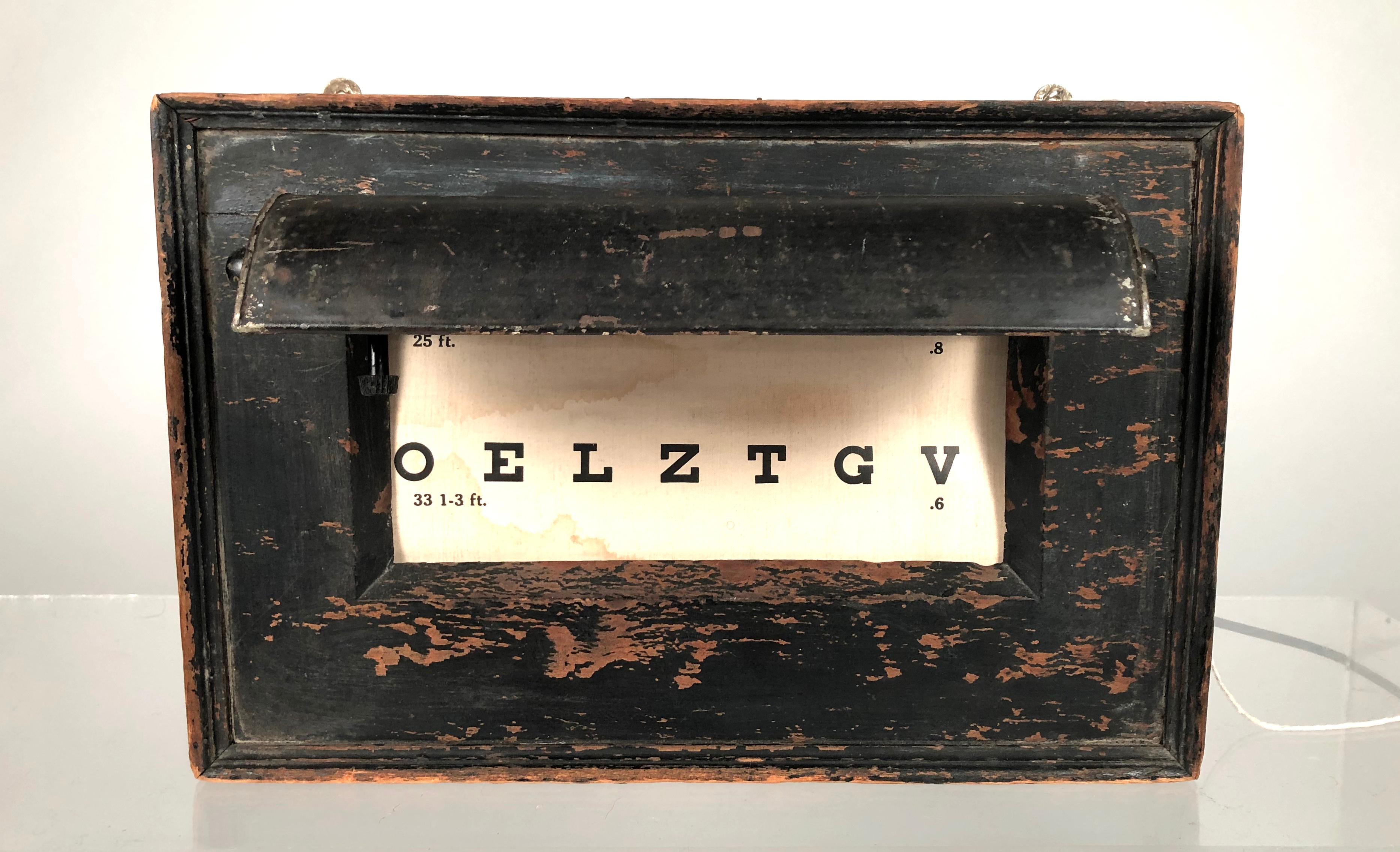 A rare, early illuminated optician's eye test chart, made by the Globe Optical Company, Boston, intended to be mounted on a wall. The rectangular black painted wooden case has decorative molding along the perimeter, a metal visor with light bulb at