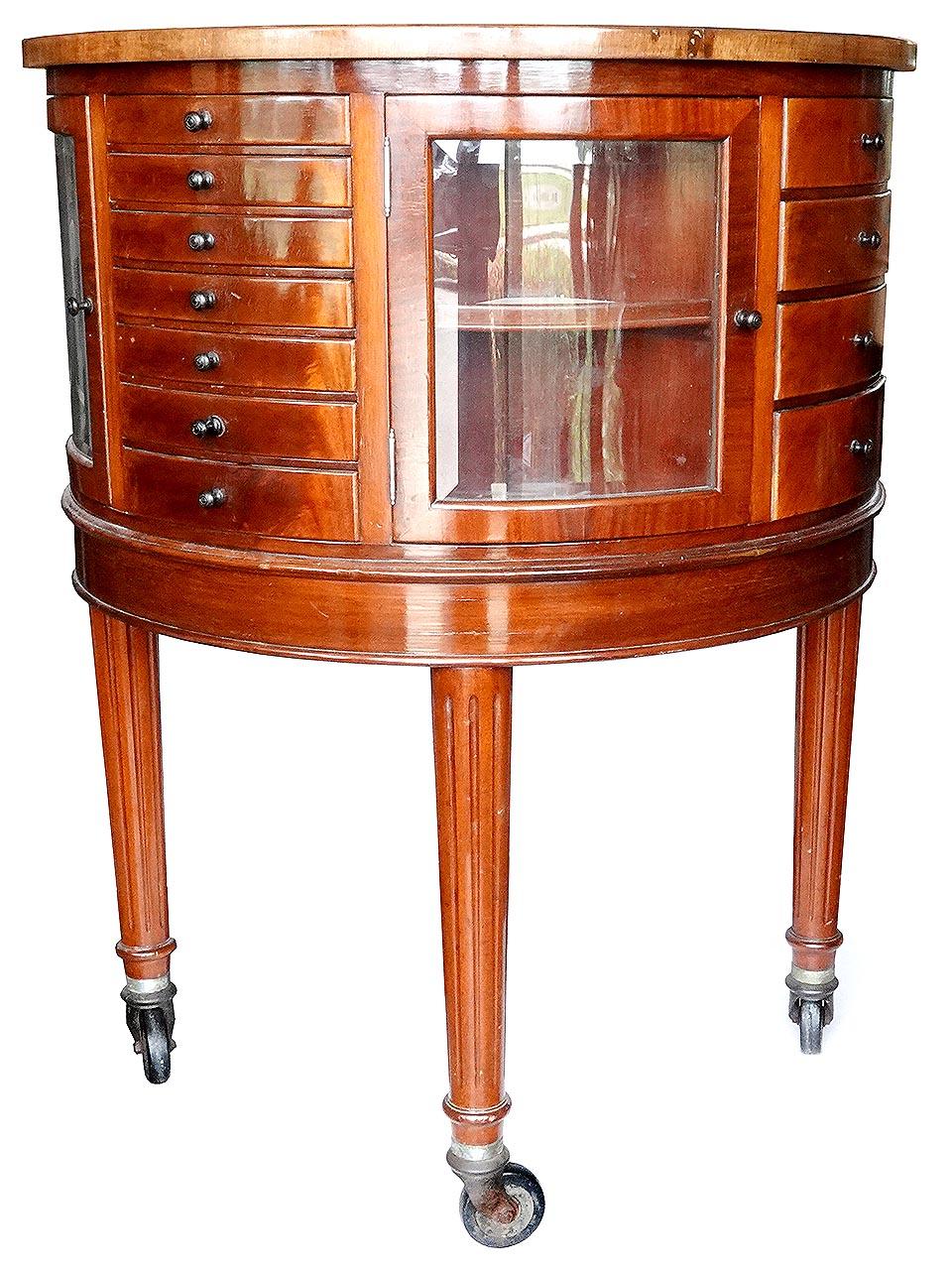 This is an antique English dental cabinet. Made from English Oak, with a circular top and fully rotating case, consisting of multiple milk glass lined drawers, glass shelves and curved bevelled glass doors, all set on turned legs, with casters. A